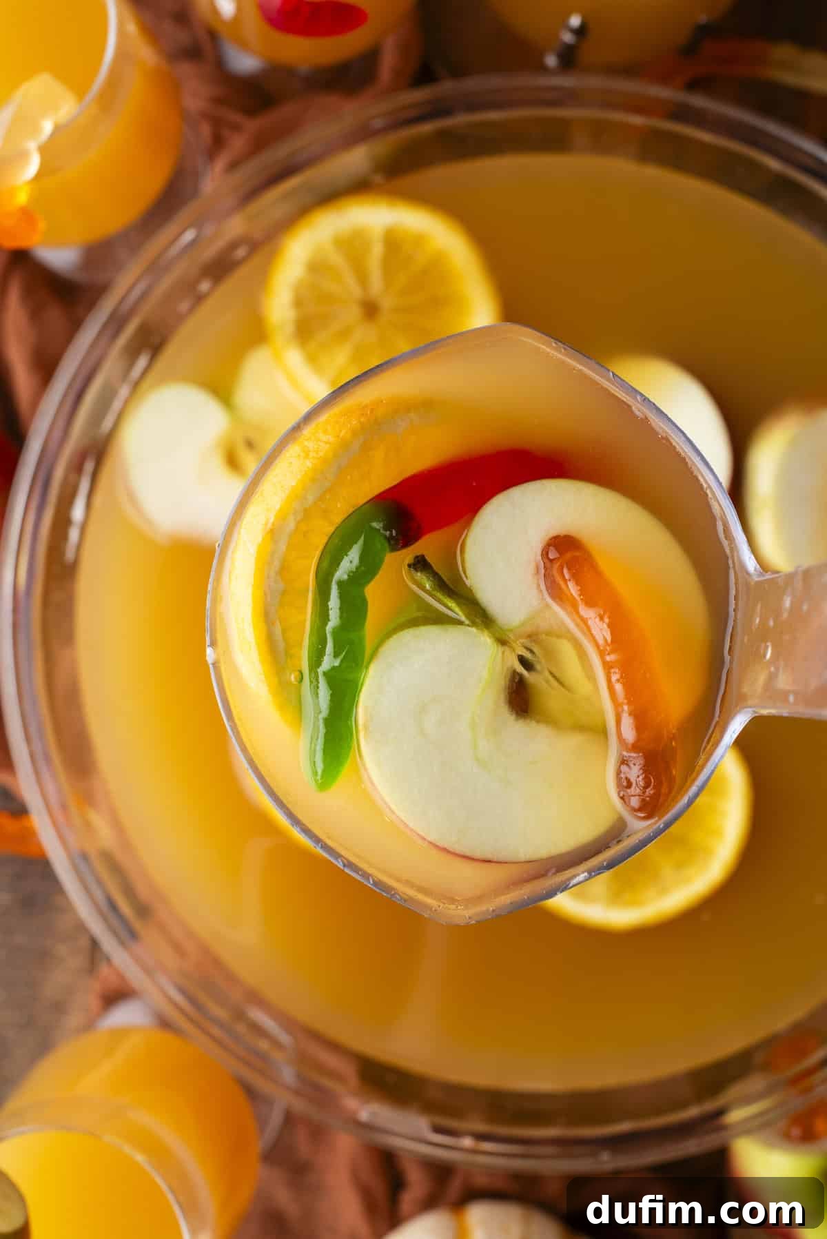 A ladle scoops vibrant orange fruit punch from a large clear bowl. The punch is brimming with colorful apple slices, orange slices, and whole green and red chili peppers for a unique touch. Several glasses filled with the same inviting drink are artfully arranged around the main bowl.