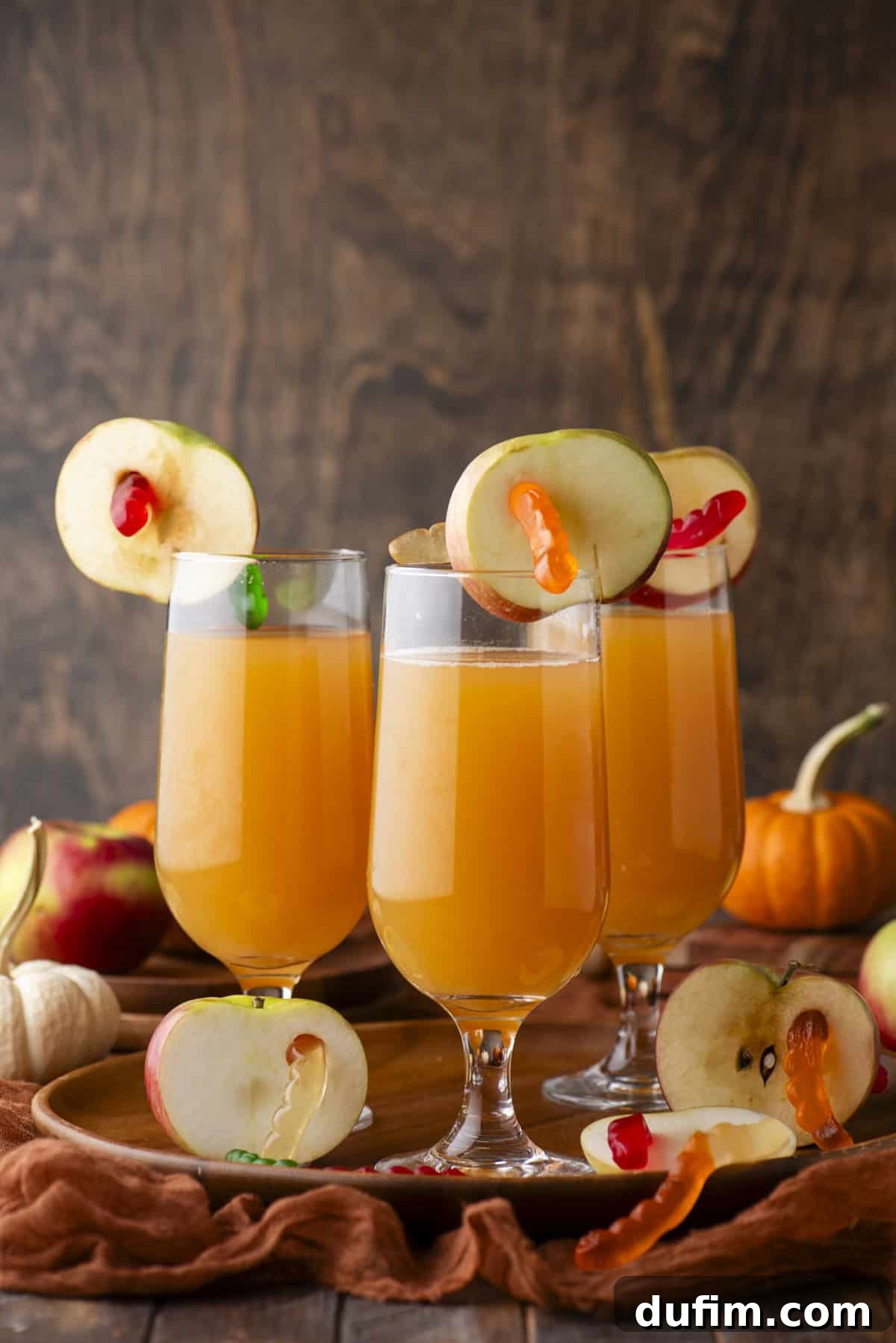 Three glasses of orange-colored drinks, elegantly garnished with apple slices and vibrant gummy worms, are arranged on a rustic wooden tray. The tray is surrounded by fresh apples, a small decorative pumpkin, and textured fabric, creating a warm and inviting autumn setting against a wooden backdrop.