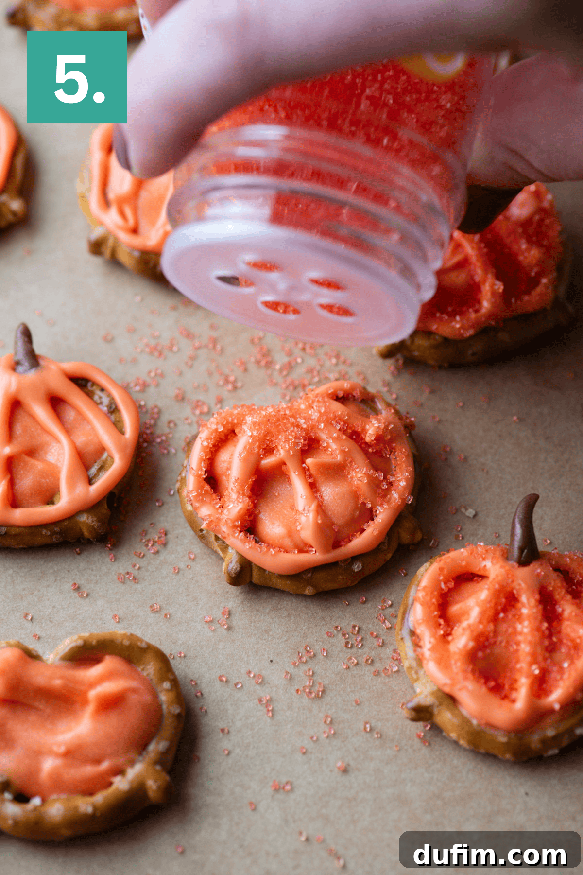 A hand sprinkles orange sugar over pretzel snacks decorated with orange icing to look like pumpkins on a sheet of parchment paper. A green box with the number 5 is in the upper left corner.
