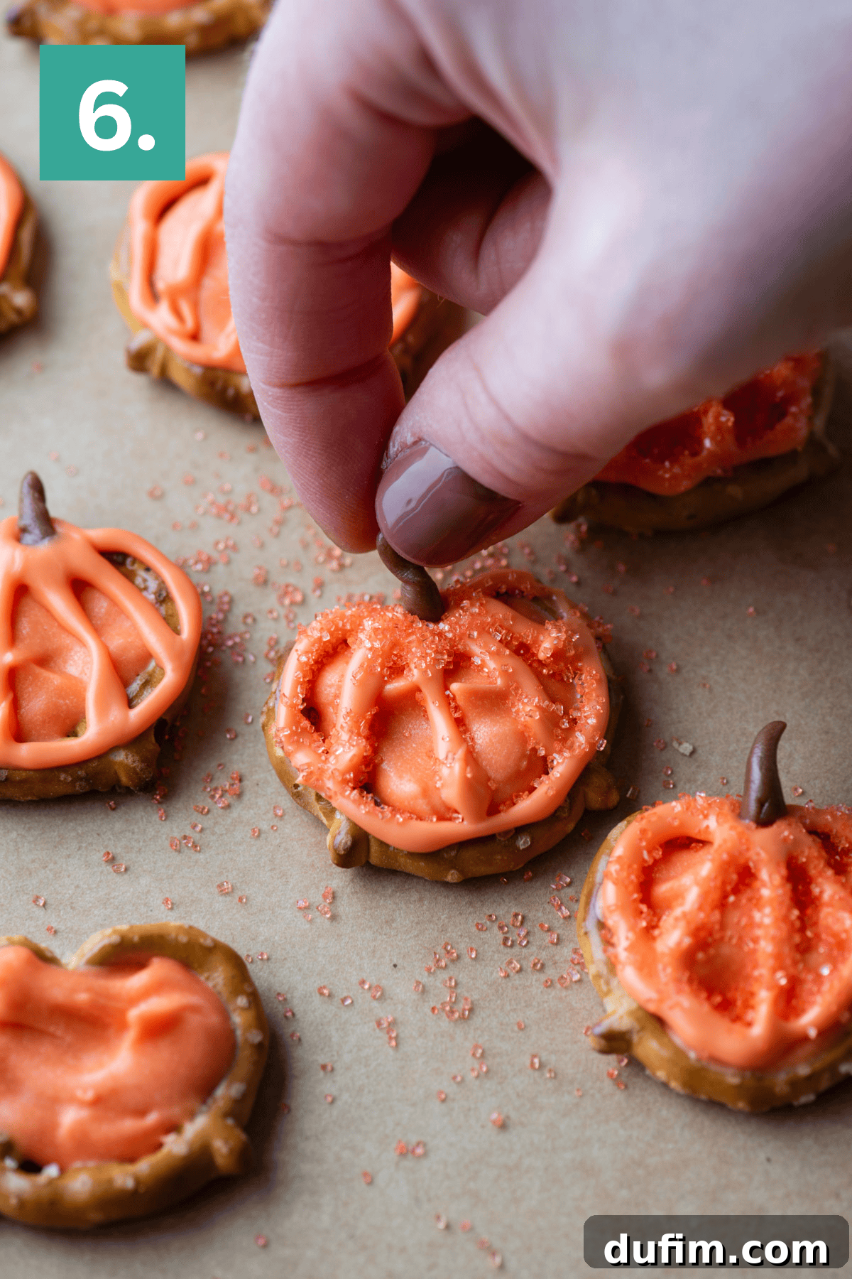 A hand places a small chocolate piece as a stem on a pretzel decorated with orange frosting to look like a pumpkin. Other similar pumpkin-shaped pretzel treats are on a sheet of parchment paper.