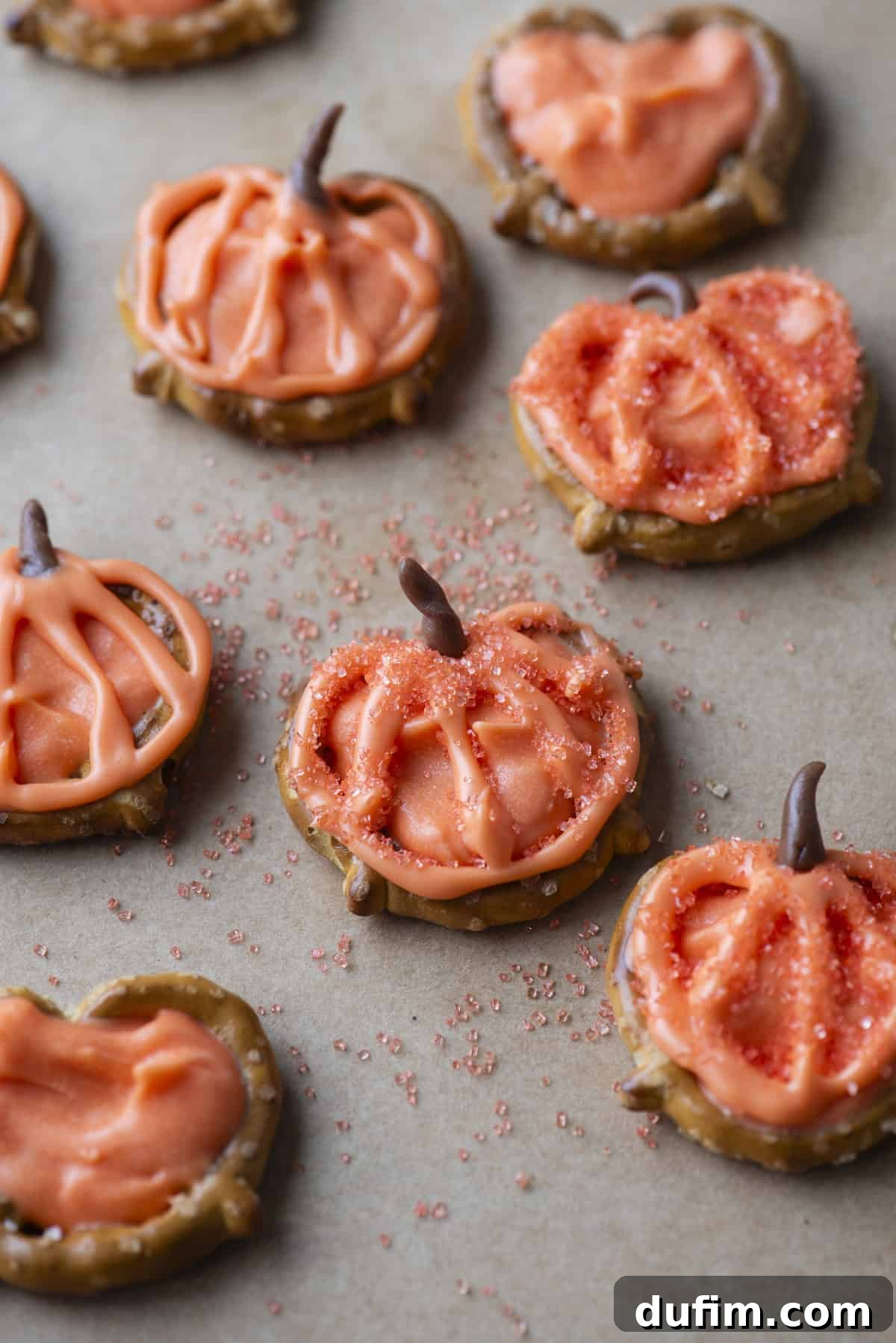 Pumpkin-shaped pretzel treats topped with orange icing and sprinkles are arranged on parchment paper. Some feature chocolate stems and decorative icing outlining pumpkin details.