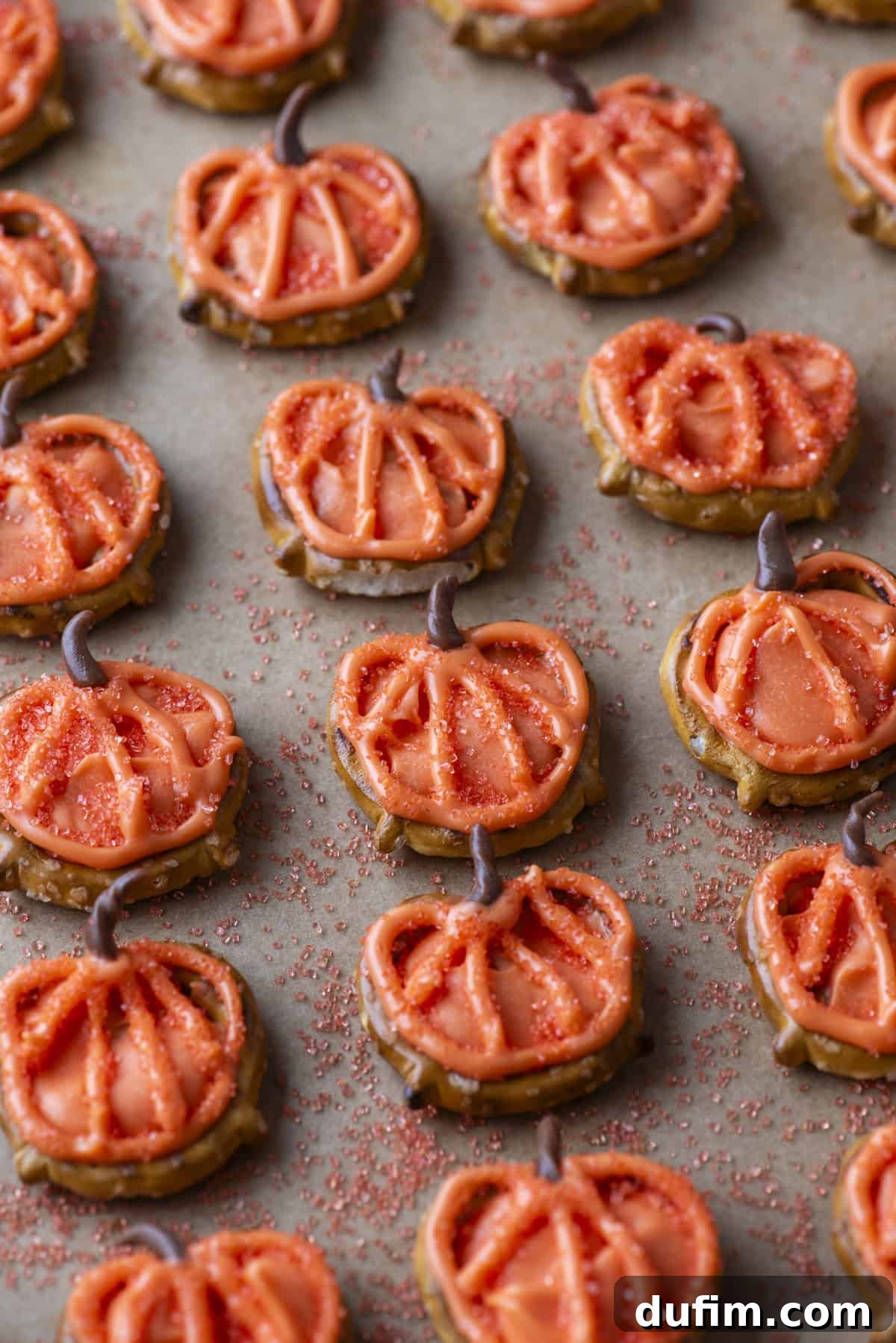 Pumpkin-shaped cookies with orange icing and red sprinkles are arranged on a baking sheet. Each cookie has a small brown stem, and red sugar crystals are scattered around them.