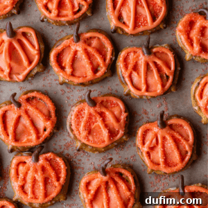 Pumpkin-shaped sugar cookies decorated with orange icing and sprinkles, arranged in rows on parchment paper. Each cookie has a chocolate stem and piped lines imitating a pumpkin’s shape.