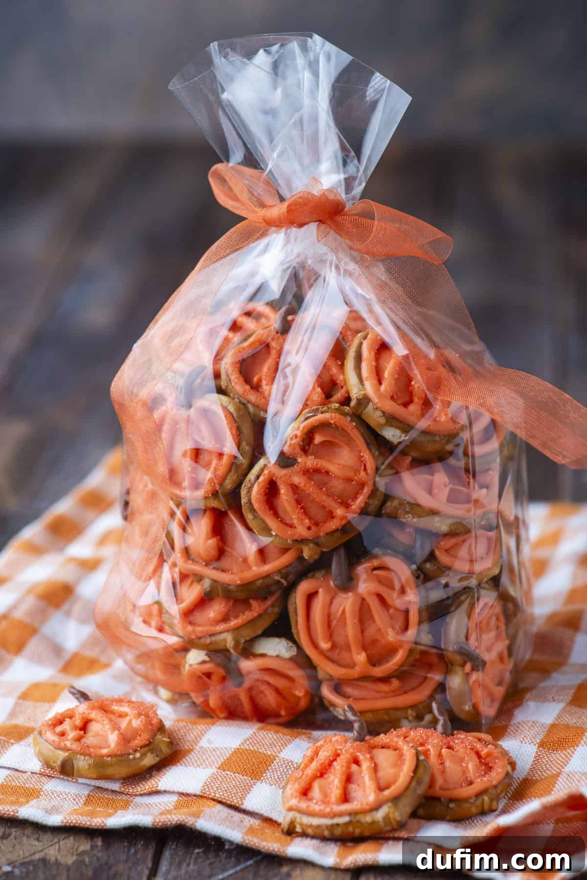 A clear plastic bag tied with an orange ribbon is filled with pretzel snacks topped with orange frosting. The bag sits on an orange-and-white checkered cloth, with a few snacks placed outside the bag.