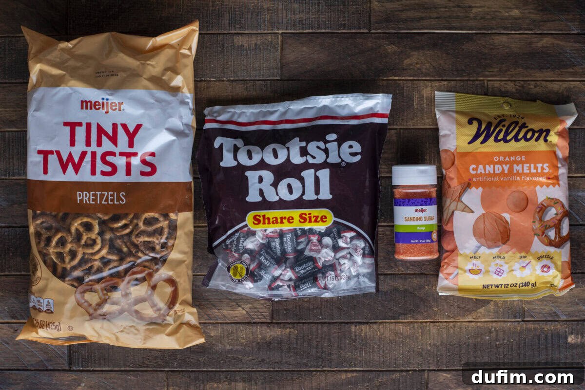 A bag of Meijer Tiny Twists pretzels, a share-size bag of Tootsie Roll candies, a container of orange sanding sugar, and a bag of Wilton orange candy melts on a wooden surface.