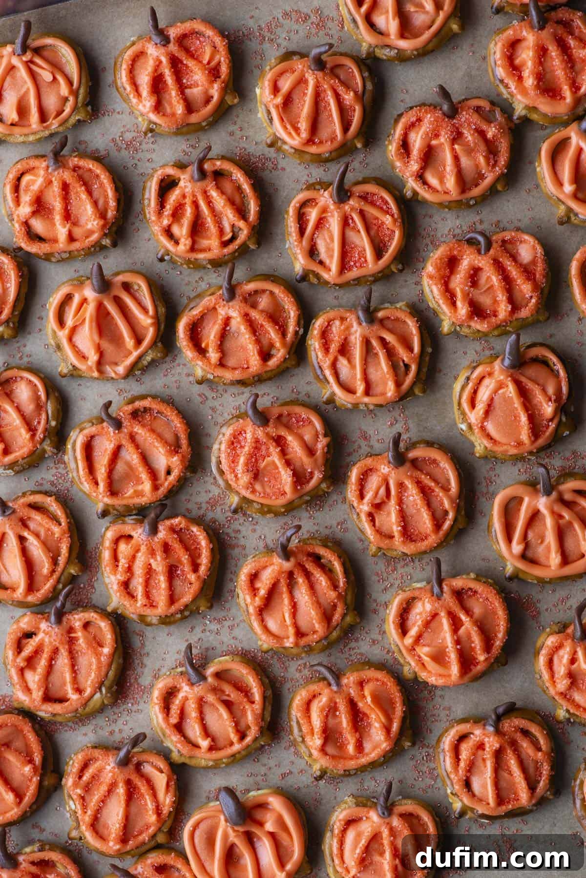 Rows of pumpkin-shaped cookies decorated with orange icing and sprinkles, each with a small chocolate stem, arranged closely together on a baking sheet.