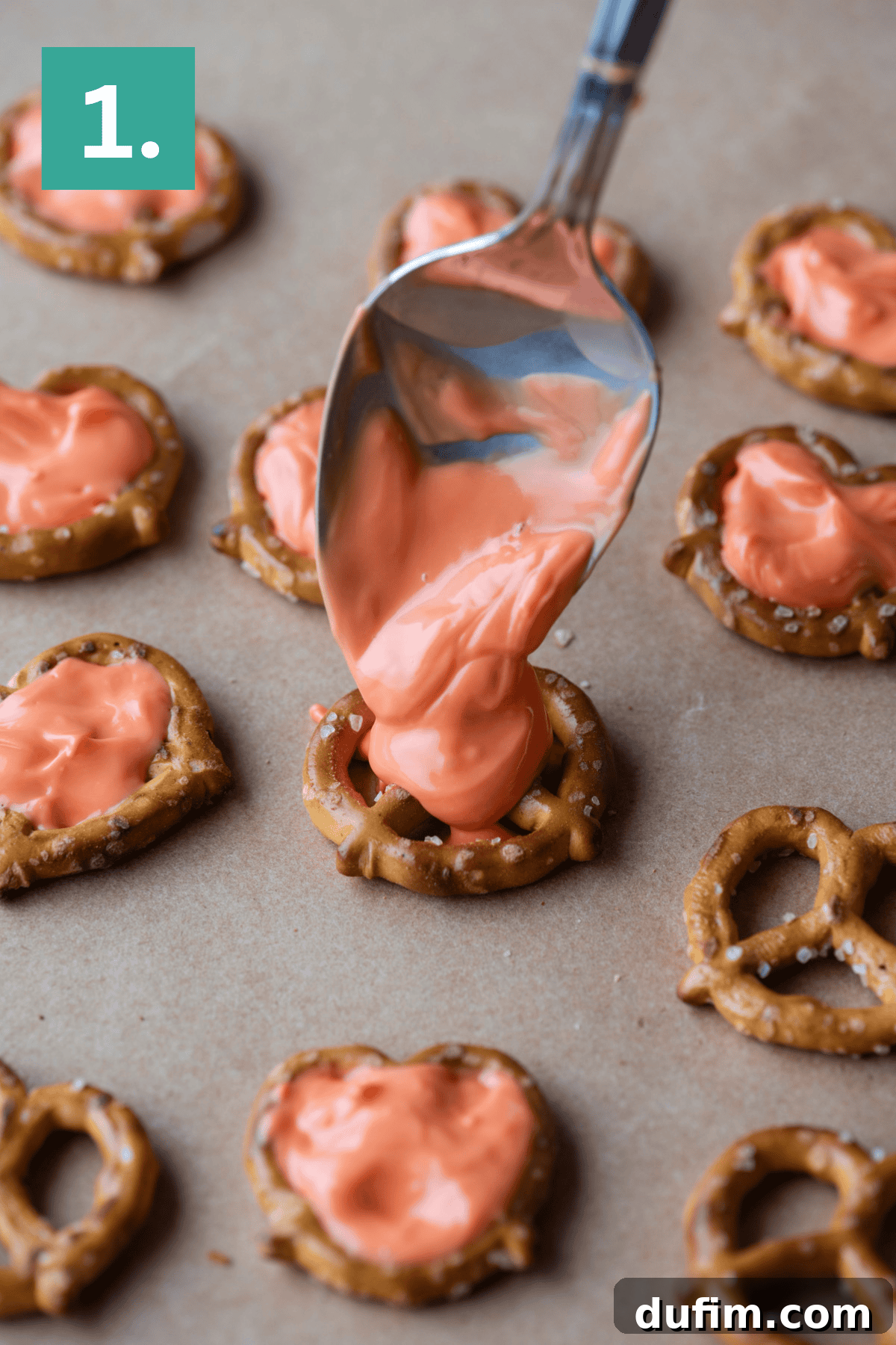 A spoon spreads melted orange-colored candy onto a small pretzel on parchment paper, surrounded by other pretzels already covered in the same candy coating. A green box with “1.” is in the top left corner.