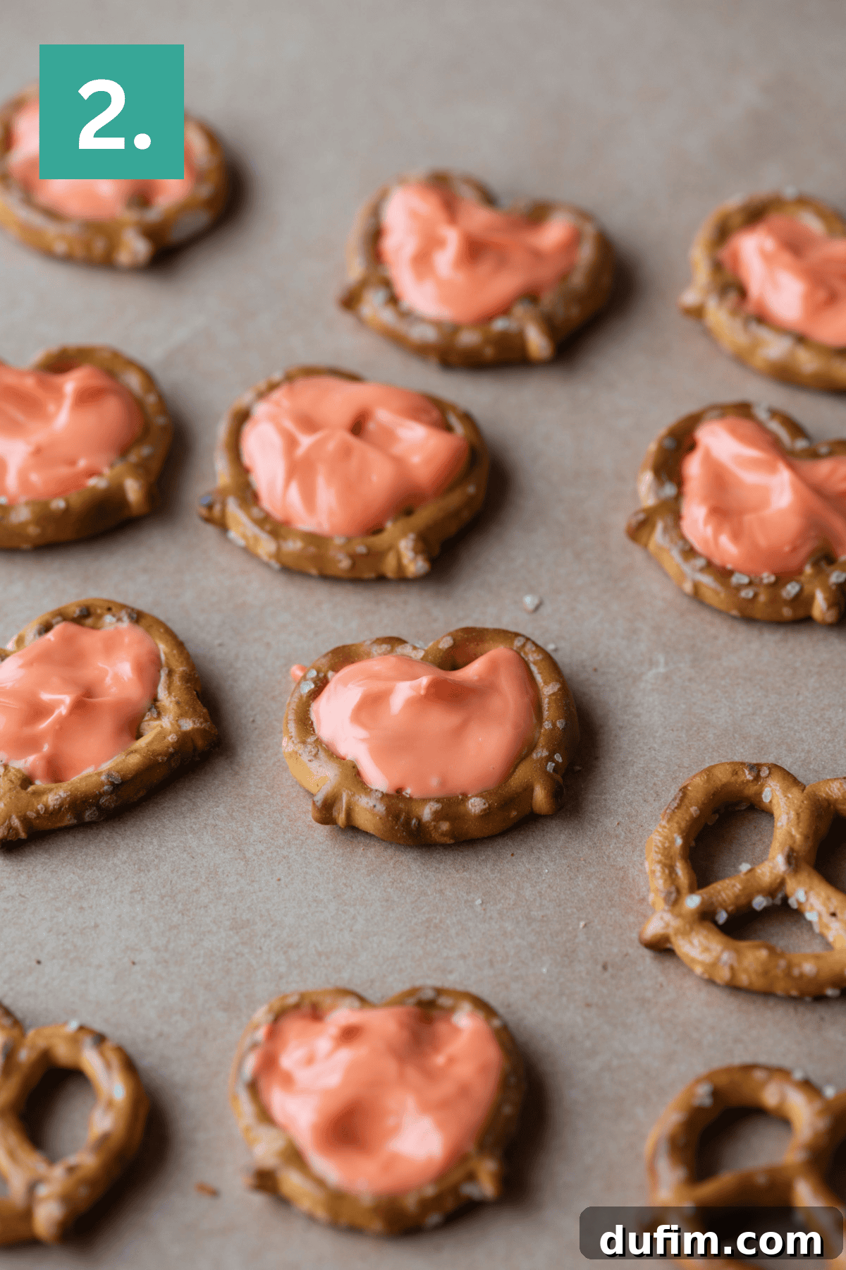 Pretzel twists spread with orange icing are arranged on a brown surface. A green square with a white number 2. appears in the top left corner.