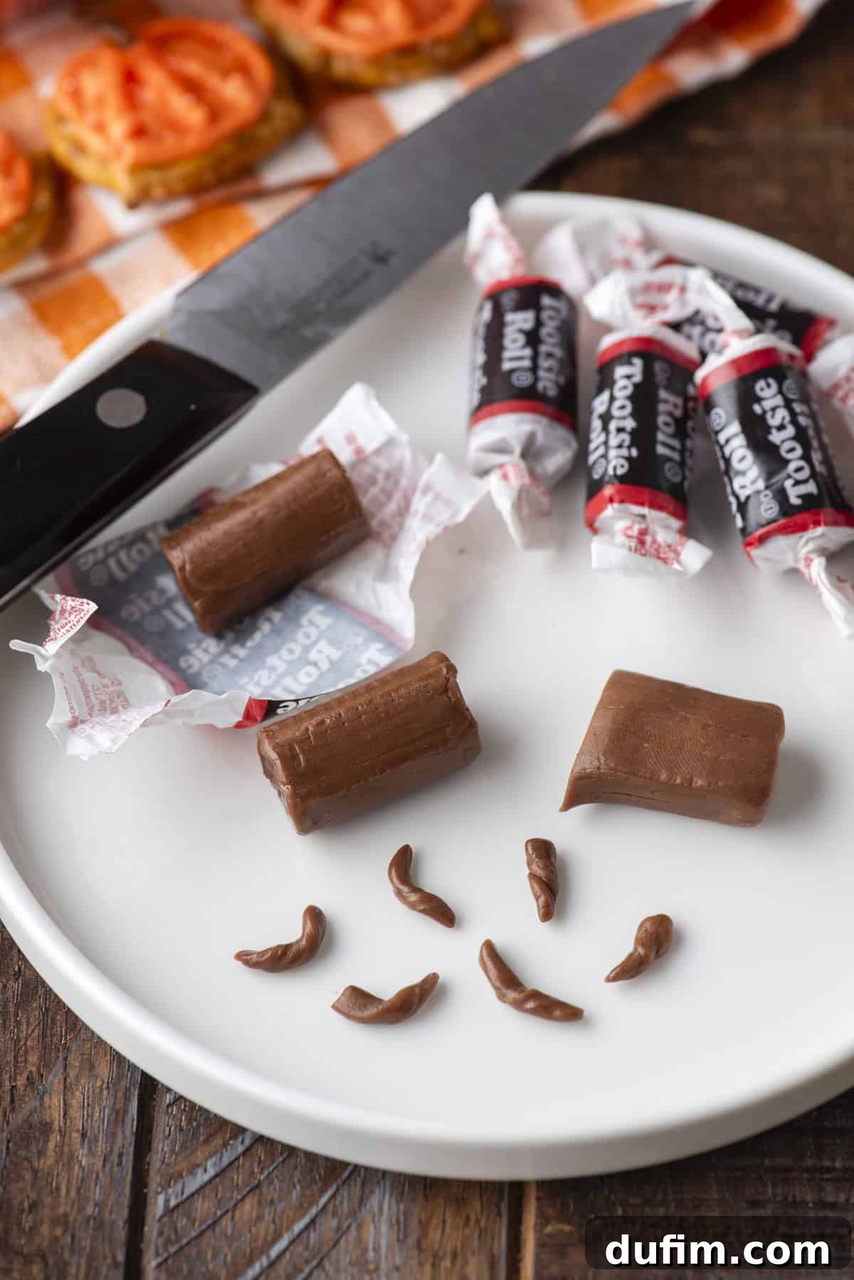 A white plate with unwrapped and wrapped Tootsie Rolls, some cut in pieces, and small pieces shaped like poop. A knife lies nearby on a checkered napkin. Everything is on a wooden table.