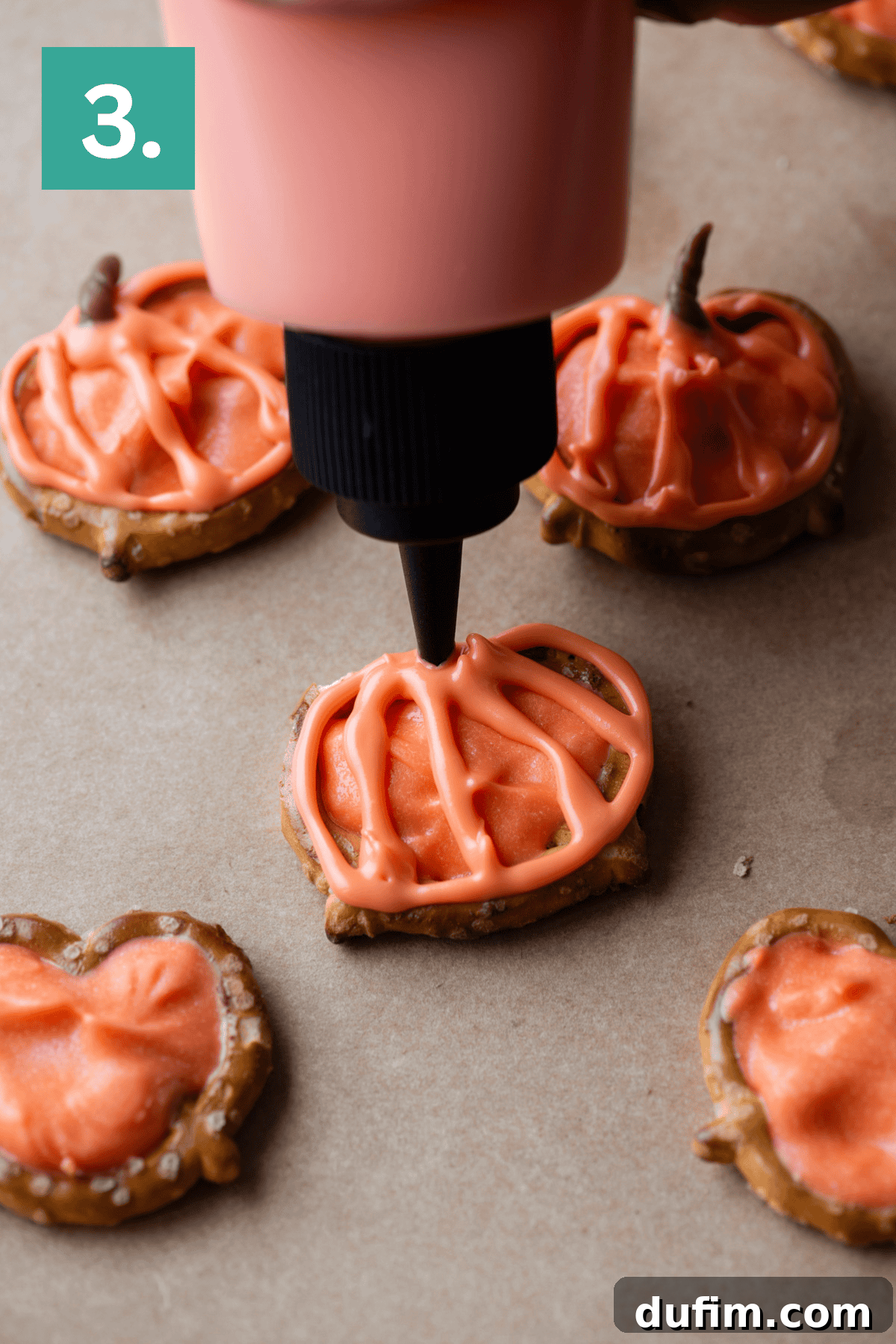 A hand uses a squeeze bottle to pipe orange icing onto a pumpkin-shaped cookie, with other iced cookies nearby on brown parchment paper. A green square with the number 3 appears in the top left corner.