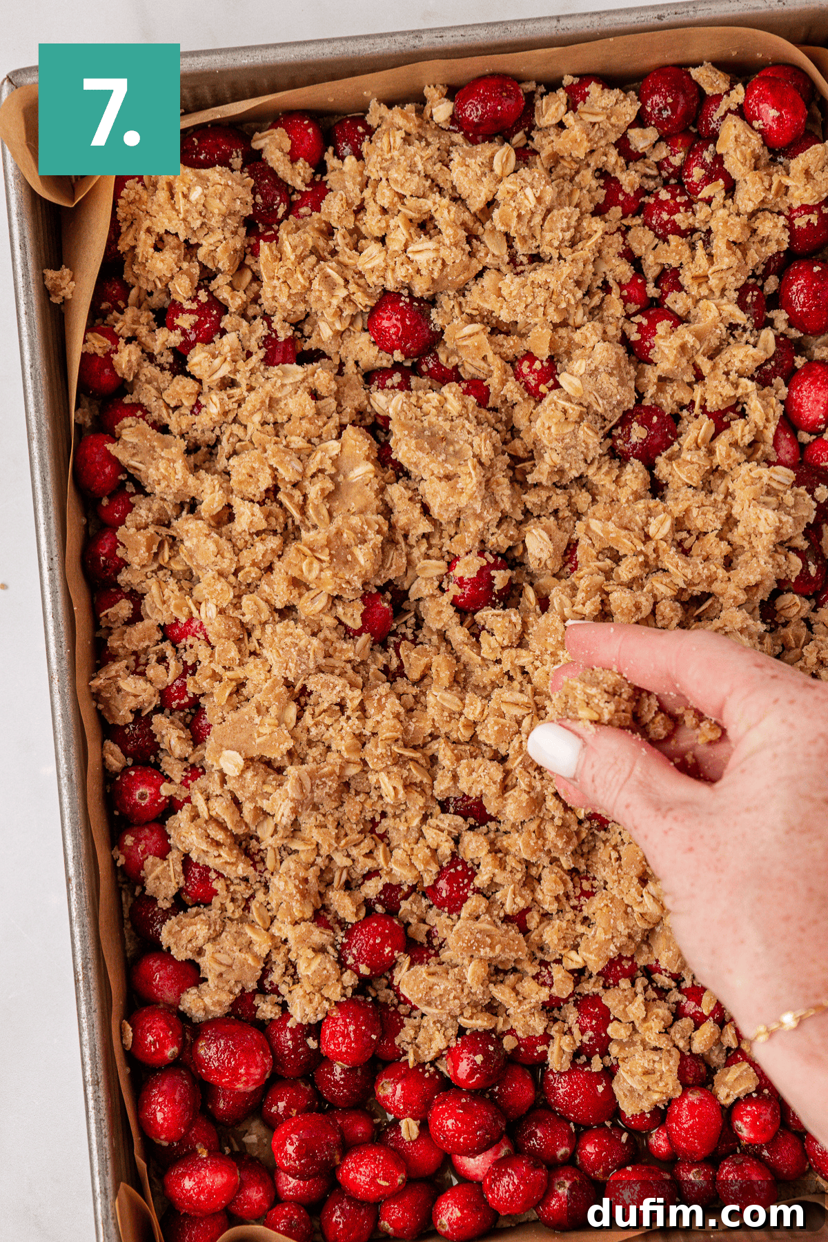 A hand sprinkles crumbly oat topping over a baking sheet filled with fresh cranberries. A green box with the number 7 is in the top left corner.