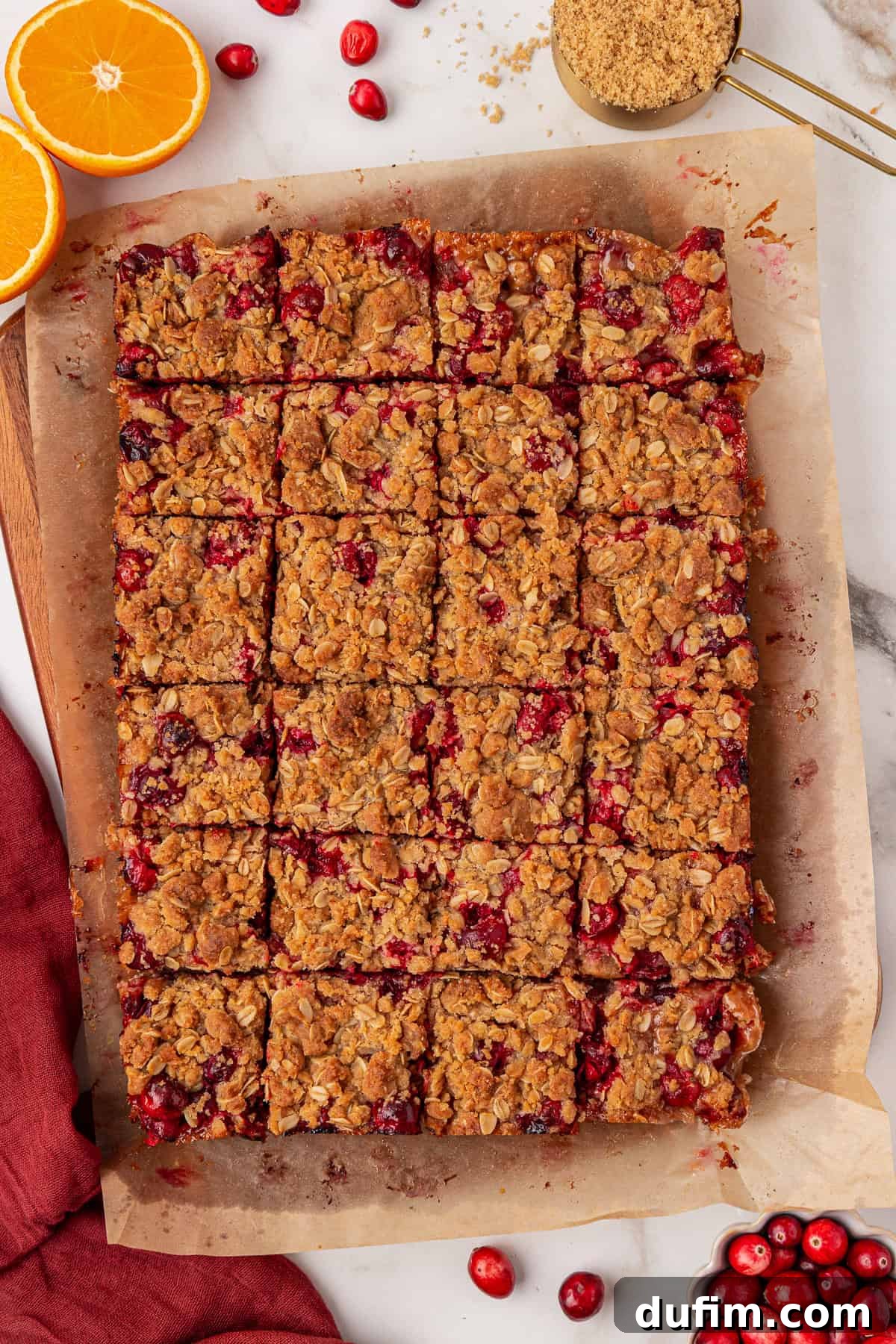 A tray of oat and cranberry bars cut into squares on parchment paper, surrounded by fresh cranberries, sliced oranges, and a measuring cup of brown sugar on a marble surface.