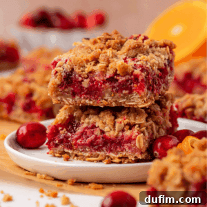 Two crumbly fruit bars with a red filling are stacked on a white plate, surrounded by fresh cranberries and orange slices. More bars and ingredients are blurred in the background.