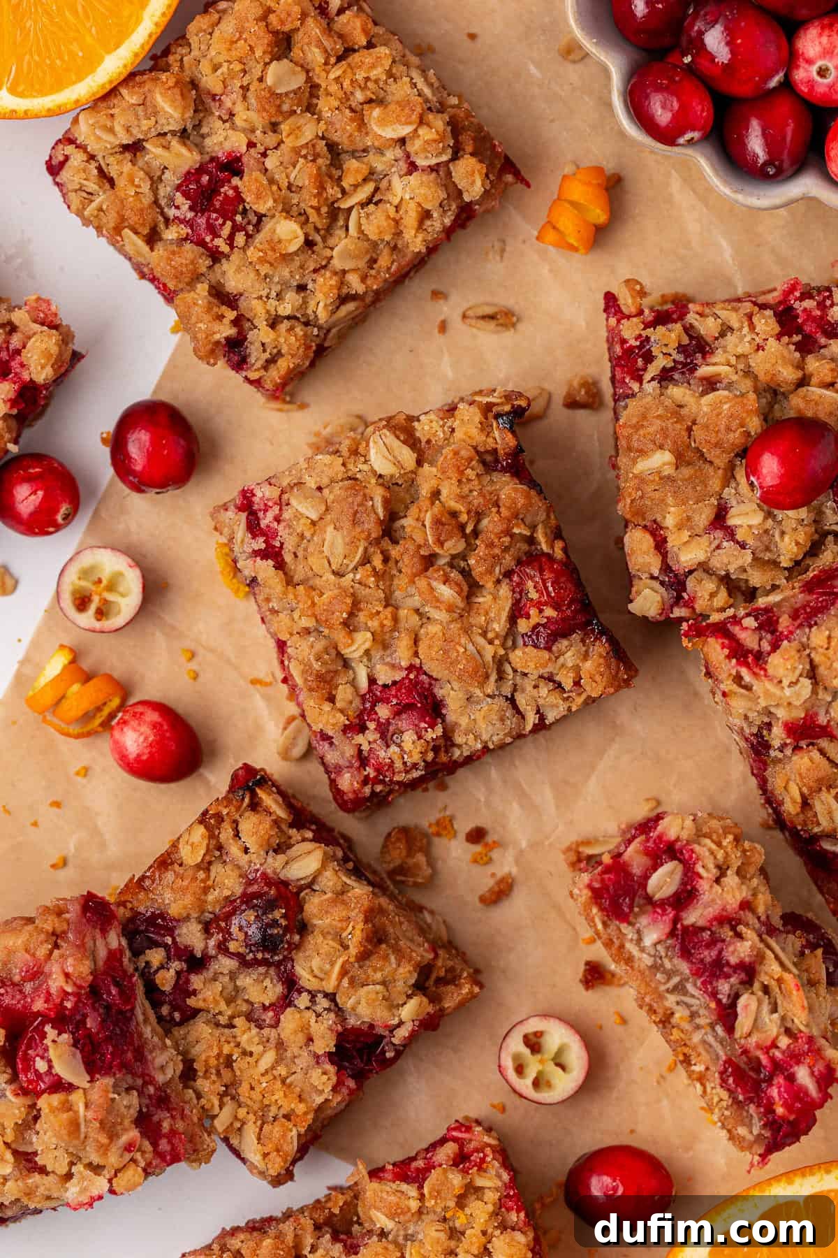 Cranberry oat crumble bars are arranged on brown parchment paper, surrounded by fresh cranberries and orange slices. The bars have a golden oat topping and visible red cranberries throughout.