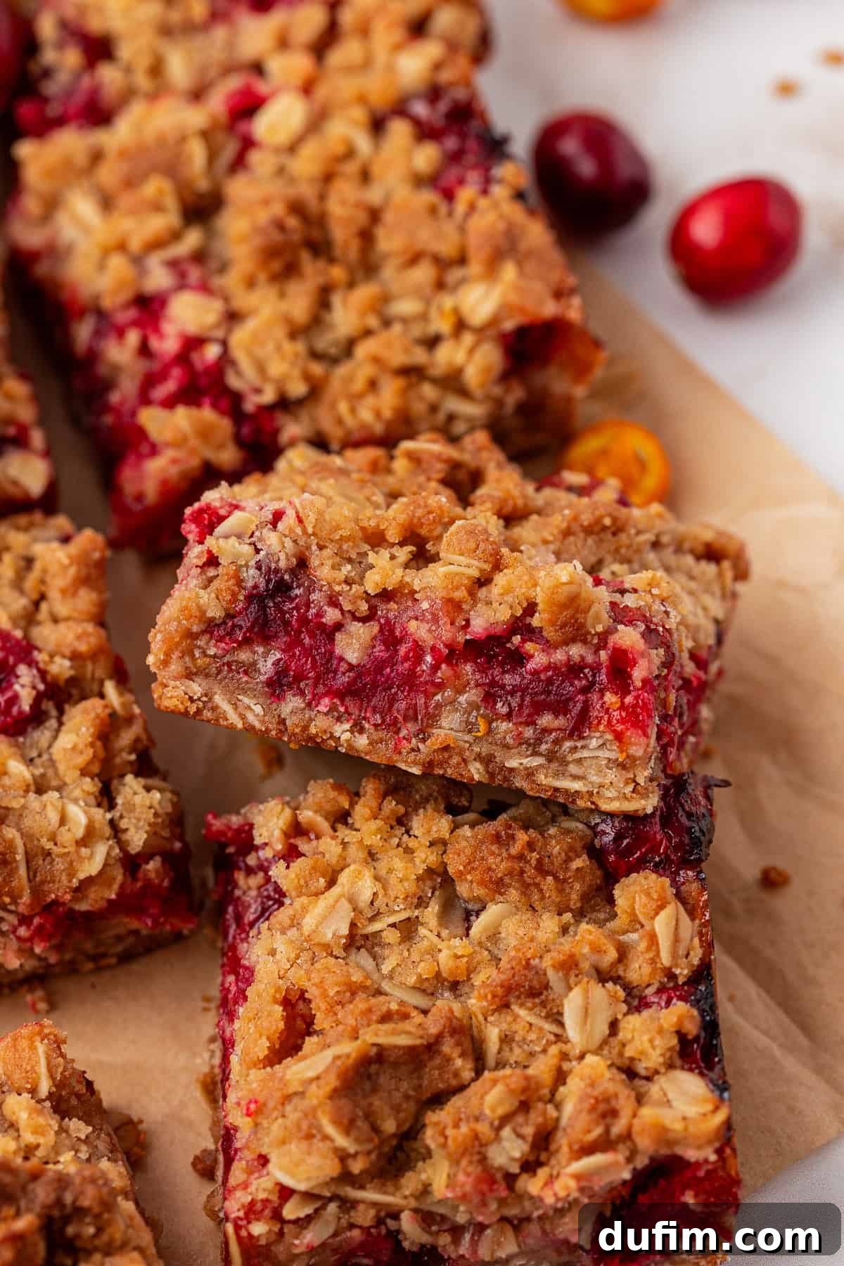 Close-up of oat crumble bars filled with a red fruit, likely cranberry filling. The bars have a golden, crumbly oat topping and are stacked on parchment paper, with a few whole cranberries nearby.