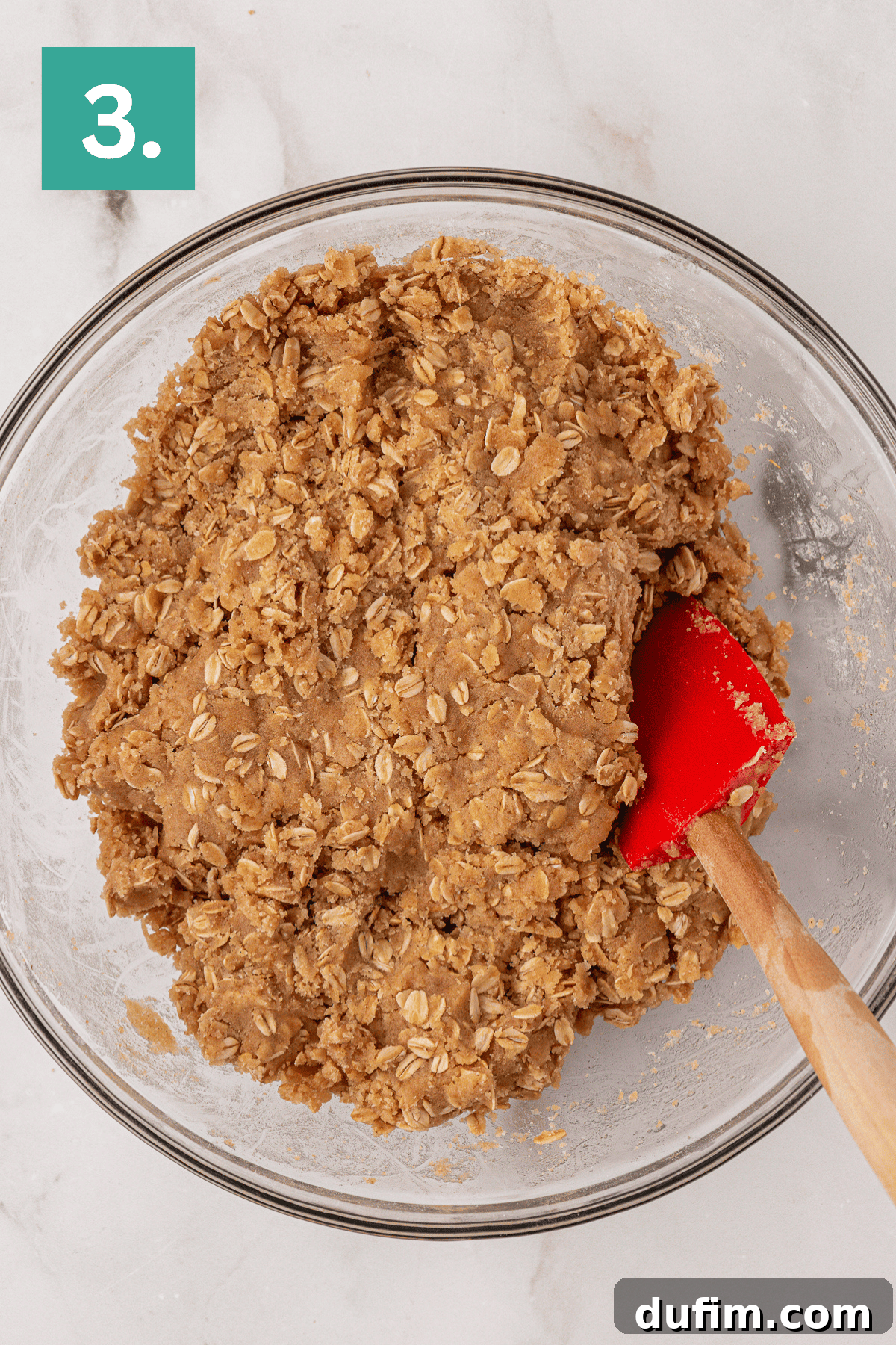 A glass bowl filled with oatmeal cookie dough is being mixed with a red spatula. A green box with the number 3 is in the top left corner. The bowl is placed on a light marble surface.