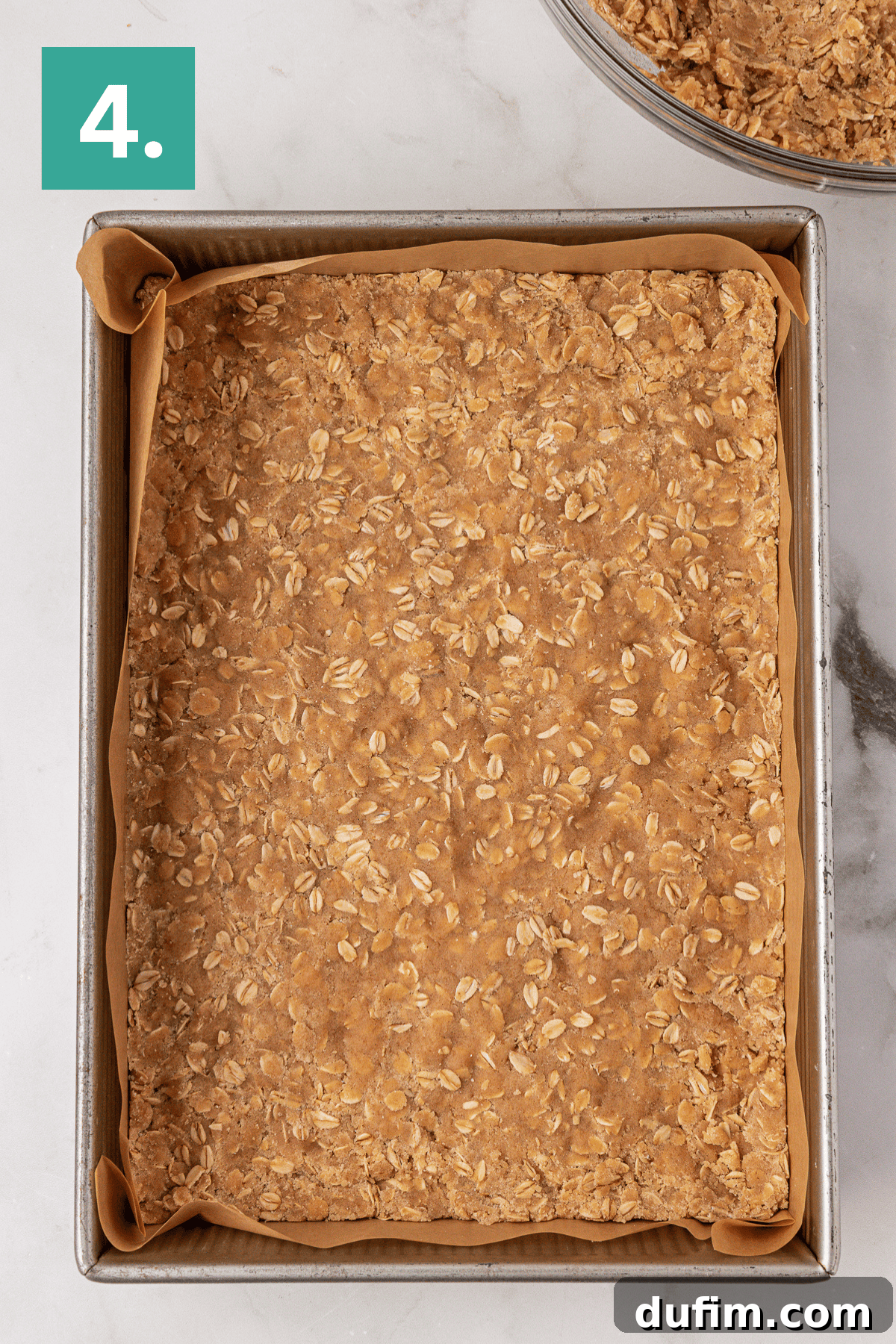 A square baking pan lined with parchment paper is filled with an unbaked oat mixture. A green box with the number 4 is in the top left corner. The pan sits on a white marble surface.