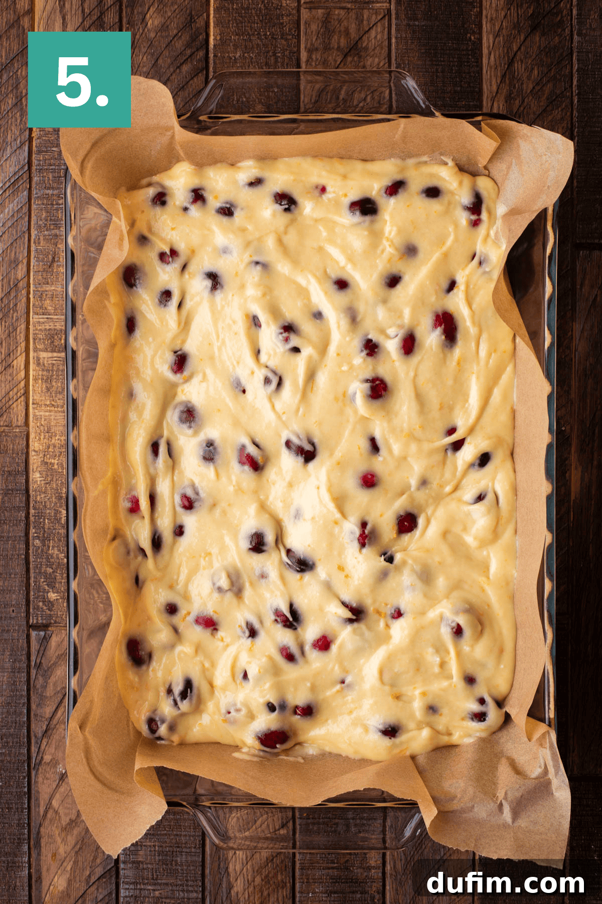 A clear glass baking dish, meticulously lined with parchment paper, holds the first even layer of raw cake batter. Scattered bright red cranberries dot the surface, ready for the next stage of layering.