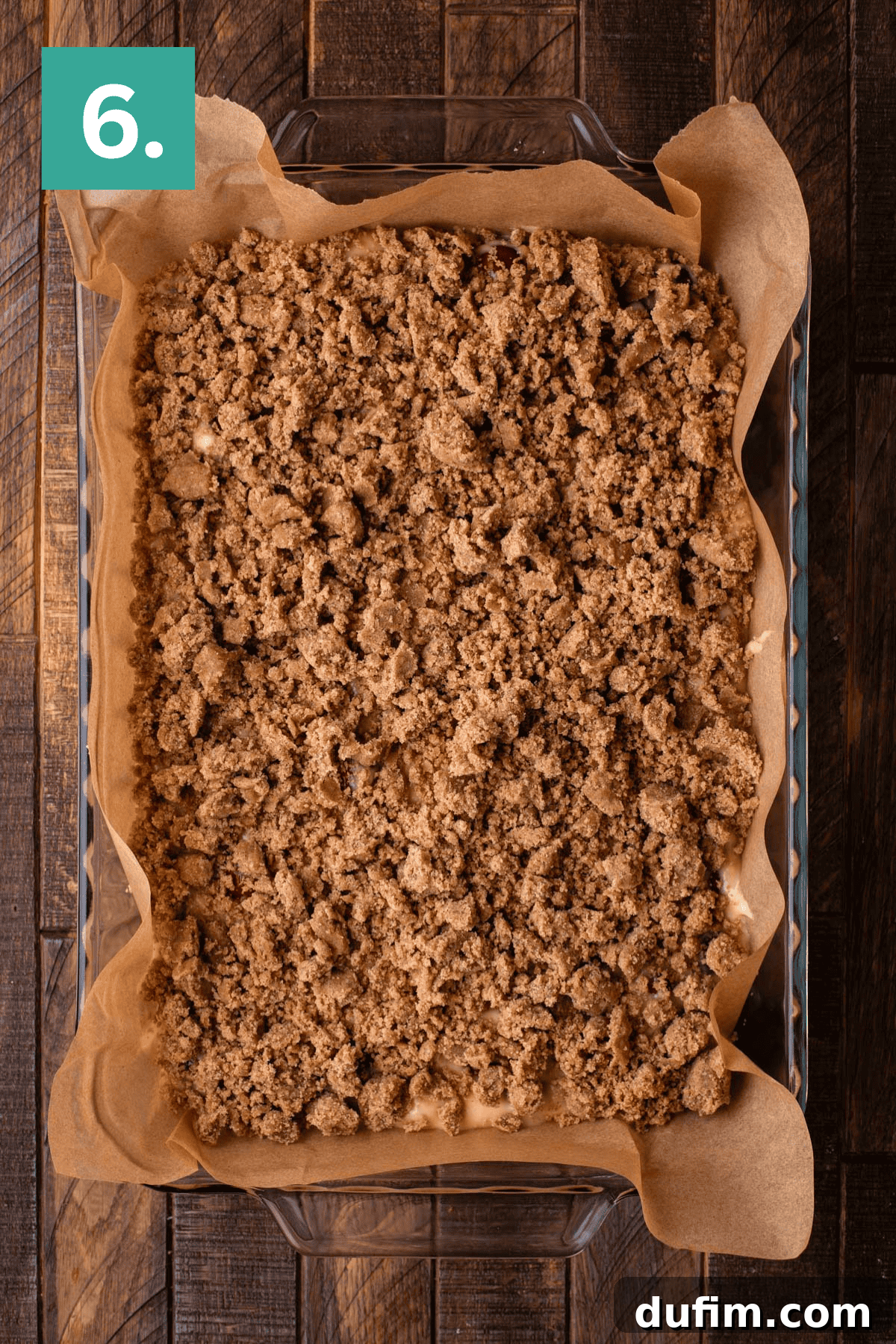 A clear glass baking dish, lined with parchment paper, showcases a large, unbaked dessert. This image highlights the second layer, featuring a generous spread of crumbly streusel topping meticulously placed over the cake batter, all set on a wooden surface.