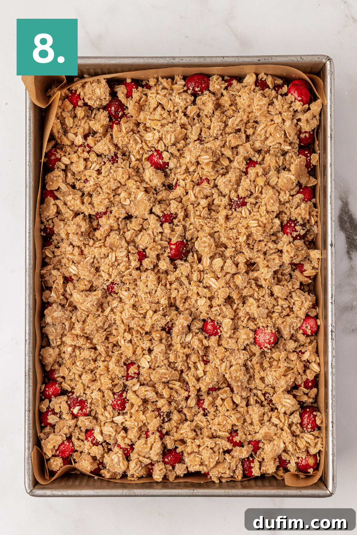 A rectangular glass baking pan, perfectly lined with parchment paper, holds an unbaked Cranberry Orange Coffee Cake. It is generously topped with a golden-brown streusel, promising a delicious texture once baked.