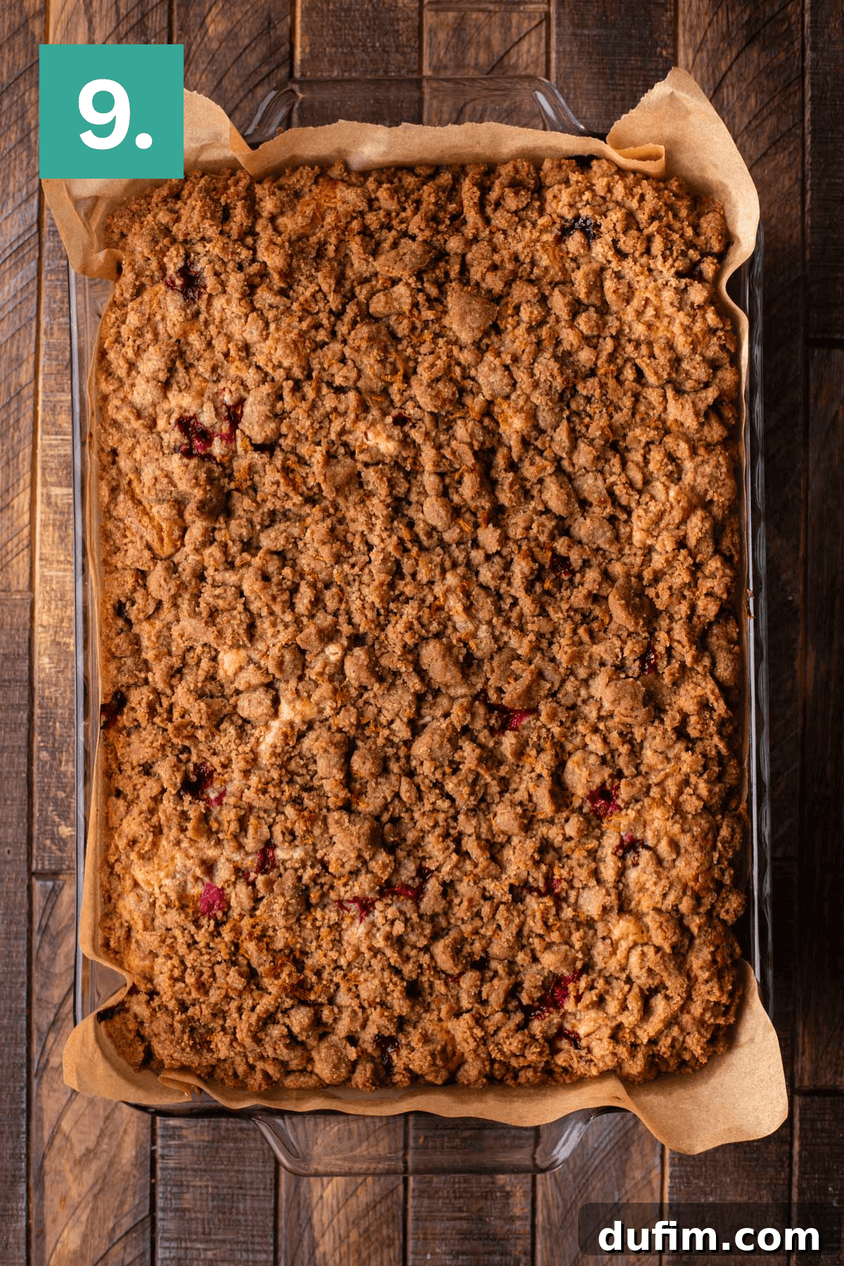 A large rectangular crumb cake, freshly baked to a golden-brown perfection with a rich streusel topping, sits invitingly in a glass baking dish lined with parchment paper. It rests on a rustic wooden surface, ready to be cooled and served.