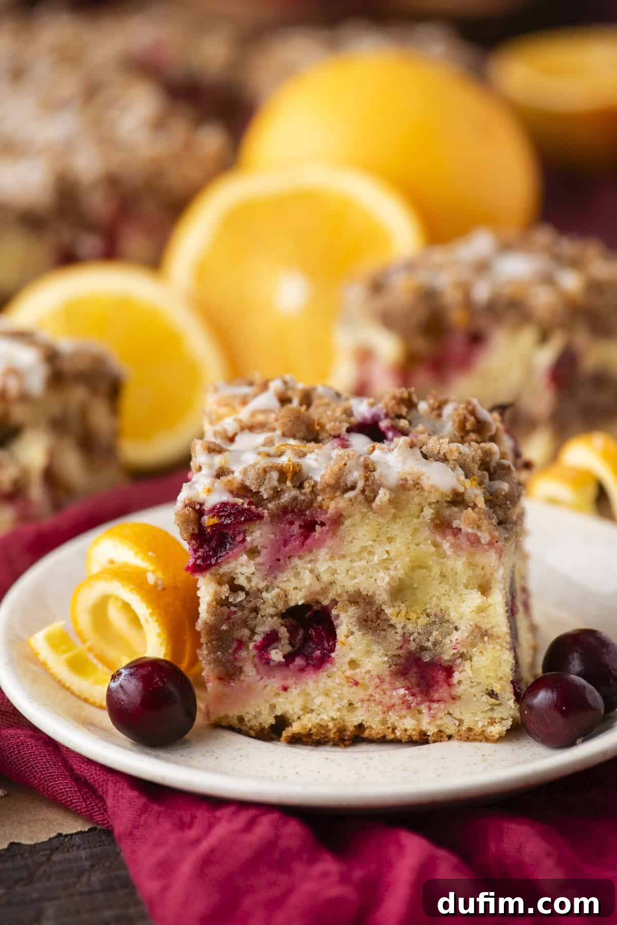 A beautifully presented slice of crumb-topped Cranberry Orange Coffee Cake, complete with visible cranberries and a delicate drizzle of orange icing. It rests on a pristine white plate, elegantly garnished with fresh orange slices and whole, vibrant cranberries. In the blurred background, whole oranges and more slices of the delicious cake hint at an abundance of this festive treat.