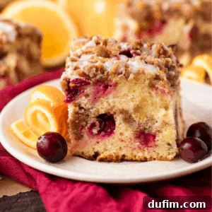 A beautifully sliced piece of Cranberry Orange Coffee Cake with crumb topping and glaze, served on a plate with fresh orange slices and cranberries.