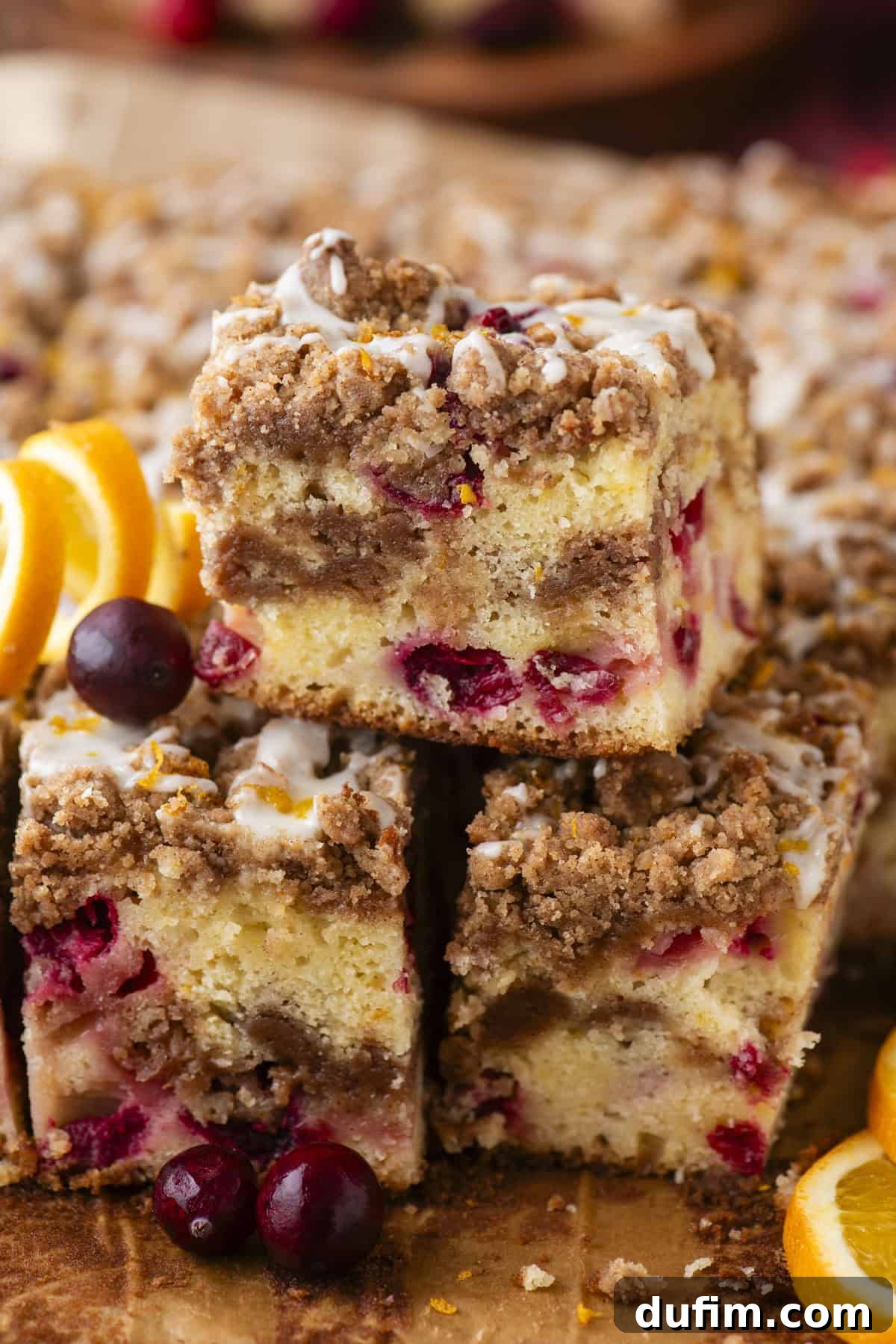 A vibrant close-up of three squares of Cranberry Orange Coffee Cake. Each piece features a rich cinnamon streusel topping and a light, citrusy glaze. Fresh, whole cranberries and bright orange slices are artfully arranged around the cake, emphasizing its fresh, seasonal ingredients.
