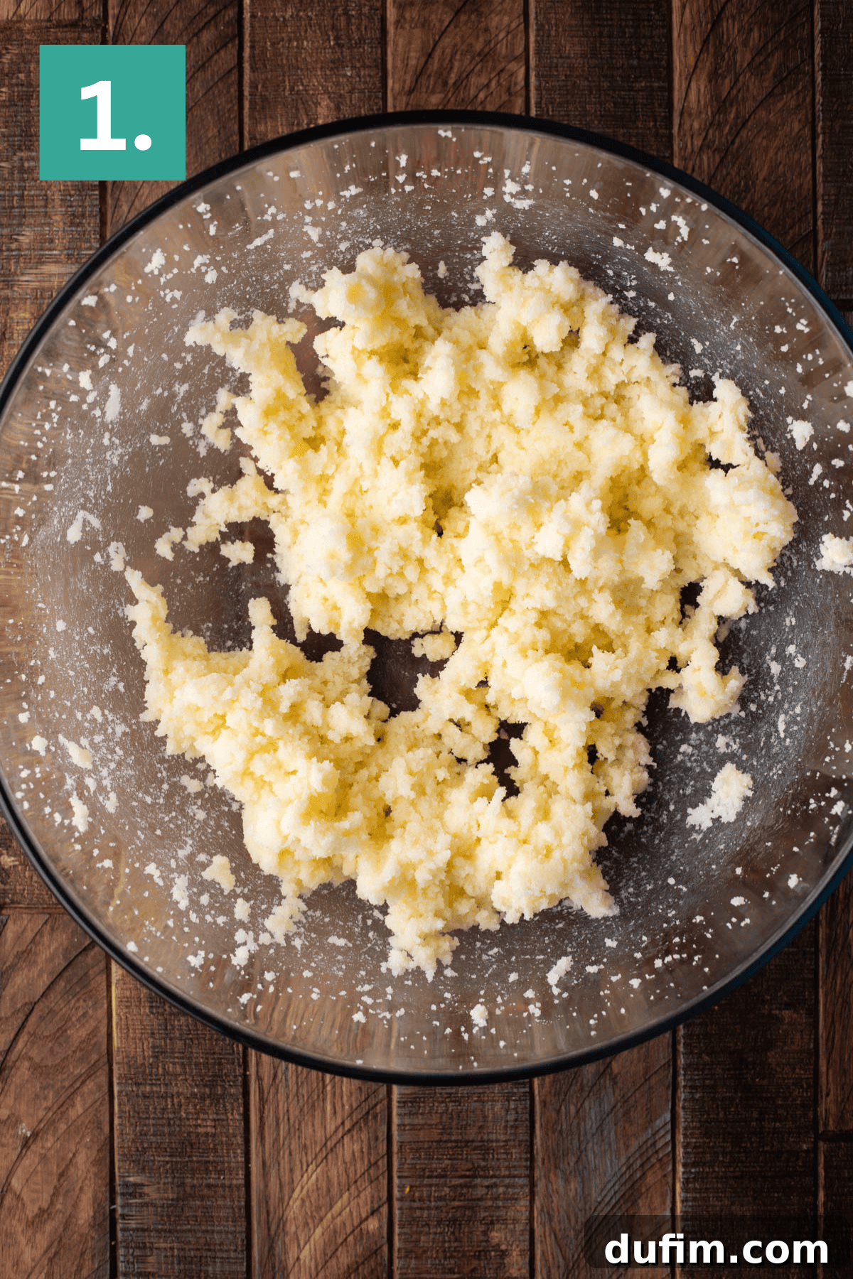 A clear glass mixing bowl contains a creamy, pale yellow mixture of butter and granulated sugar, beautifully light and fluffy after being creamed. The bowl rests on a warm wooden surface, ready for the next baking step.