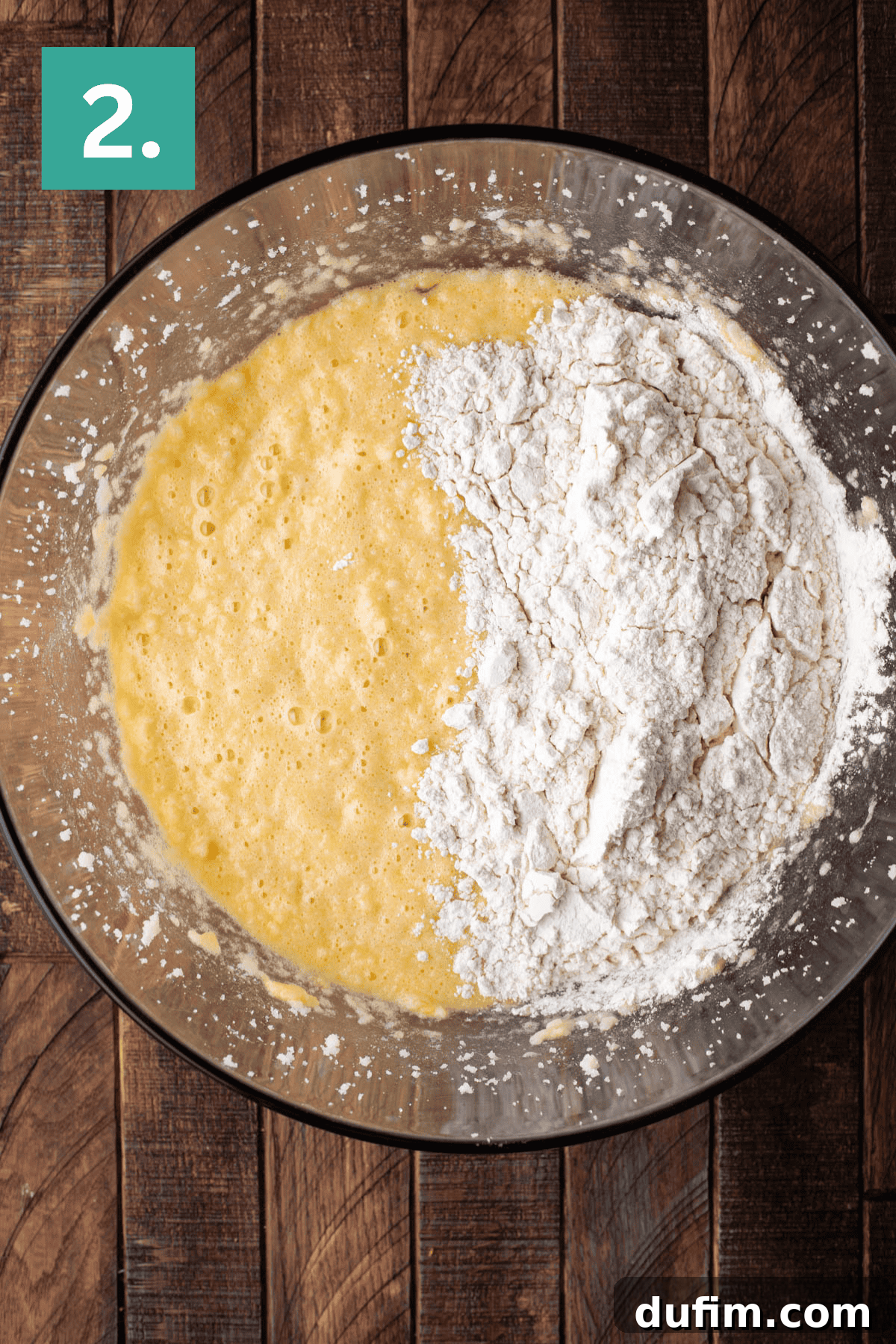 A clear glass bowl on a wooden surface holds a pale yellow cake batter, lightly dusted with white flour on top. This image captures the moment dry ingredients are about to be mixed into the wet batter, indicating the crucial next step in the baking process.