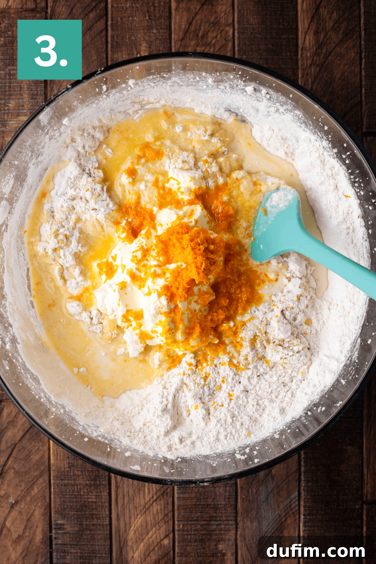 A clear glass mixing bowl on a wooden surface displays cake batter being actively stirred with a teal spatula. Visible additions include flour, creamy sour cream, and bright orange zest, indicating the aromatic citrus infusion.