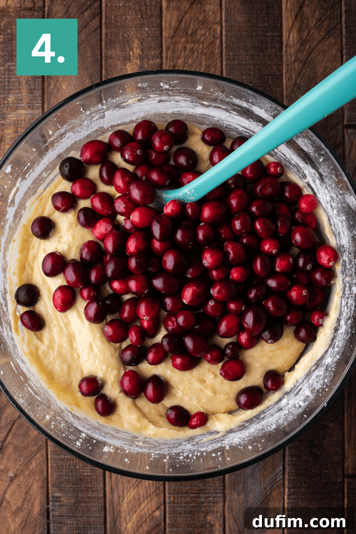 A clear glass bowl contains a rich, fluffy cake batter, with vibrant fresh cranberries being gently folded in with a teal spatula. The bowl sits on a wooden surface, showcasing the integration of the fruit into the batter.