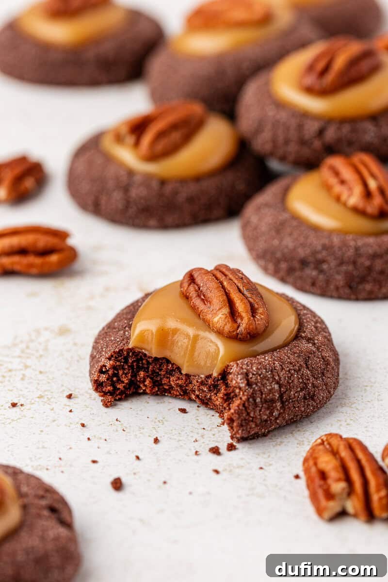 Caramel Pecan Turtle Thumbprints 2 Chocolate thumbprint cookies topped with caramel and pecan halves are arranged on a white surface; one cookie in front has a bite taken out, showing a chewy texture inside. Scattered pecans and cookie crumbs surround the cookies.