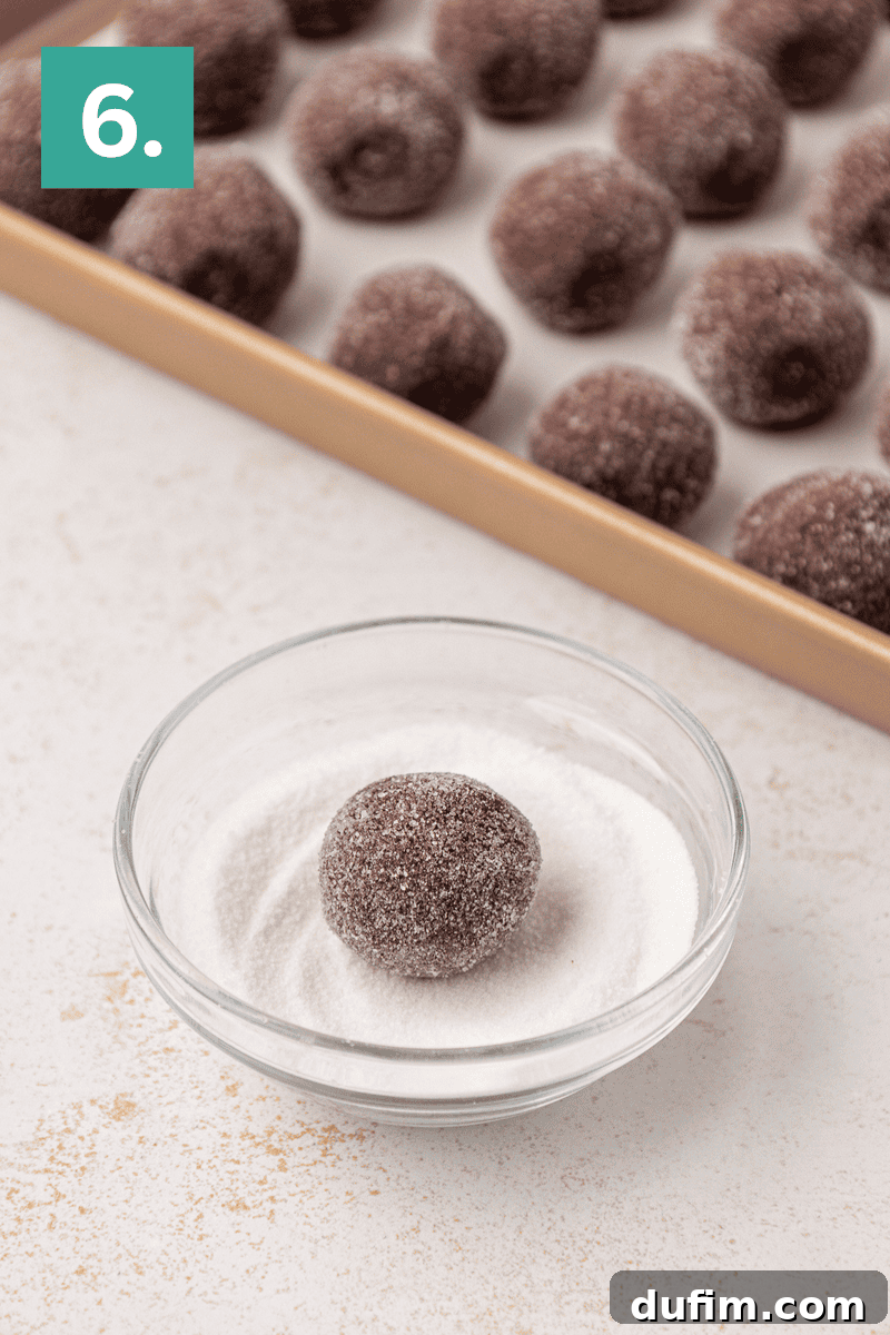 Caramel Pecan Turtle Thumbprints 11 A chocolate dough ball is being coated in sugar in a glass bowl, with more sugar-coated dough balls on a baking sheet in the background. A green box with the number 6 is in the top left corner.
