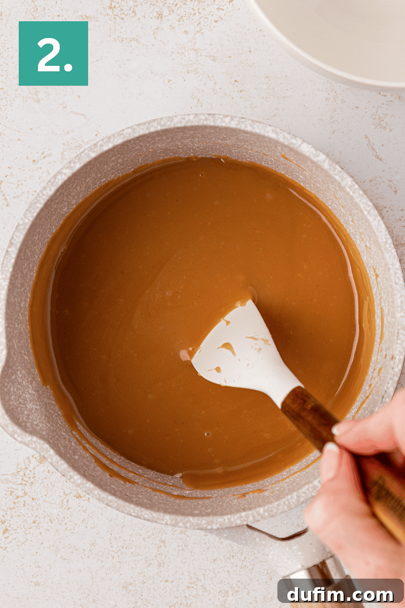 Caramel Pecan Turtle Thumbprints 15 A hand stirs smooth caramel sauce with a white spatula in a light-colored saucepan. A green box with the number 2 is in the upper left corner.