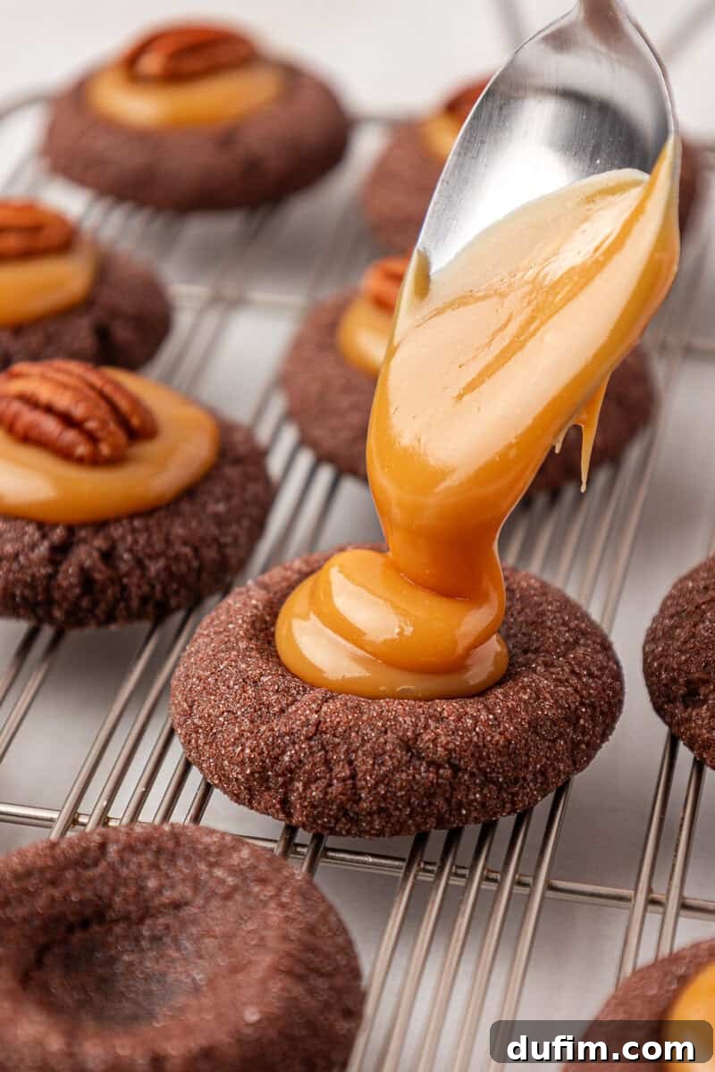 Caramel Pecan Turtle Thumbprints 16 A spoon drizzles caramel onto a round chocolate cookie topped with a pecan, while similar cookies cool on a wire rack in the background.