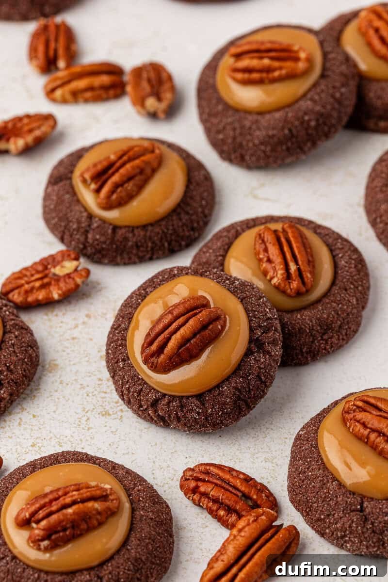 Caramel Pecan Turtle Thumbprints 17 Round chocolate cookies topped with a glossy caramel layer and a whole pecan, scattered on a white surface with some loose pecans around them.