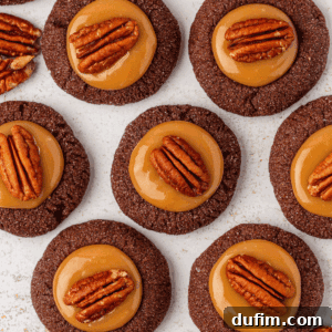 Chocolate cookies topped with a layer of caramel and a pecan half, arranged neatly on a white surface. The cookies have a glossy caramel center, and the pecans add a decorative touch.