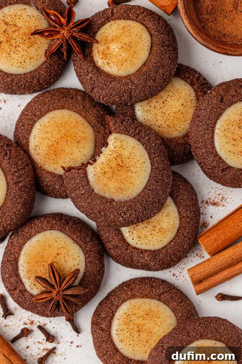 A close-up of chocolate cookies with creamy centers, sprinkled with cinnamon, surrounded by cinnamon sticks, star anise, and cloves on a white surface. One cookie has a bite taken out of it.