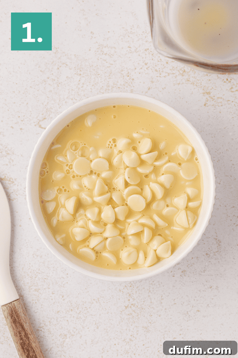 White bowl filled with yellow batter and topped with white chocolate chips, placed on a light-colored surface next to a spatula. A green box with the number “1.” is in the upper left corner.