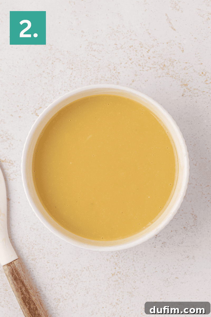 A bowl of smooth, yellow soup sits on a light-colored surface next to a utensil with a wooden handle. A green square with the number 2. is in the top left corner.