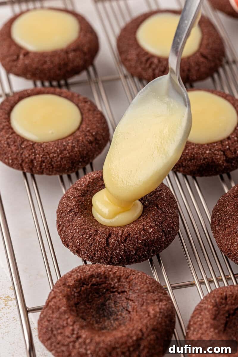 A spoon is filling the center of a chocolate thumbprint cookie with creamy yellow filling on a wire cooling rack, surrounded by other similar cookies.
