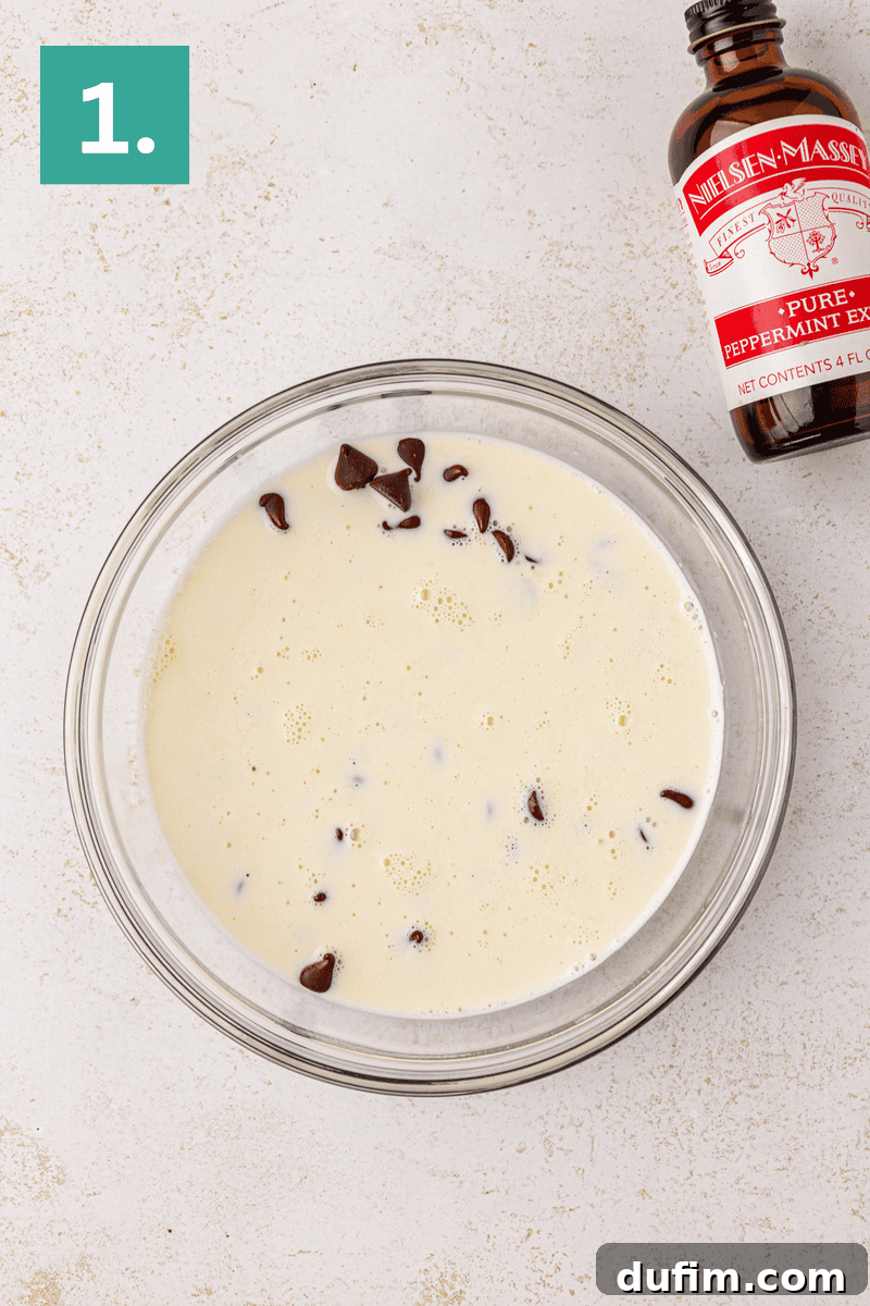Peppermint Kissed Chocolate Thumbprint Cookies 15 A glass bowl with milk and chocolate chips sits on a light surface. To the upper right is a bottle labeled “Peppermint Extract.” A green square with the number 1 appears in the top left corner.