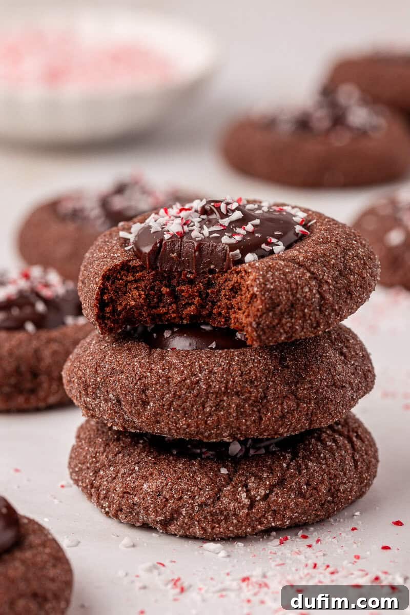 Peppermint Kissed Chocolate Thumbprint Cookies 17 A stack of three chocolate cookies topped with chocolate and crushed peppermint, with the top cookie showing a bite taken out. More cookies and peppermint sprinkles are scattered in the background.