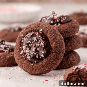 A stack of chocolate thumbprint cookies filled with glossy chocolate ganache and sprinkled with crushed peppermint pieces, with more cookies and peppermint crumbs in the background.