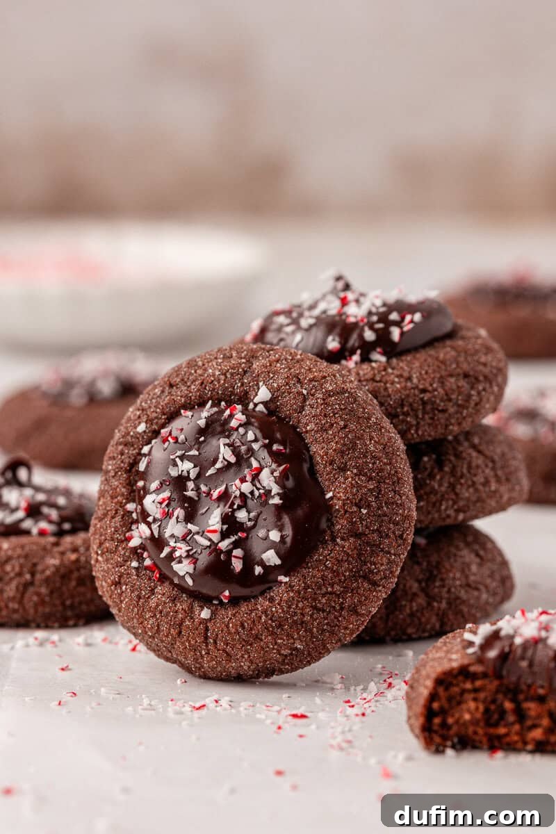 Peppermint Kissed Chocolate Thumbprint Cookies 4 A stack of chocolate thumbprint cookies filled with melted chocolate and sprinkled with crushed peppermint, with one cookie leaning against the stack and more cookies blurred in the background.