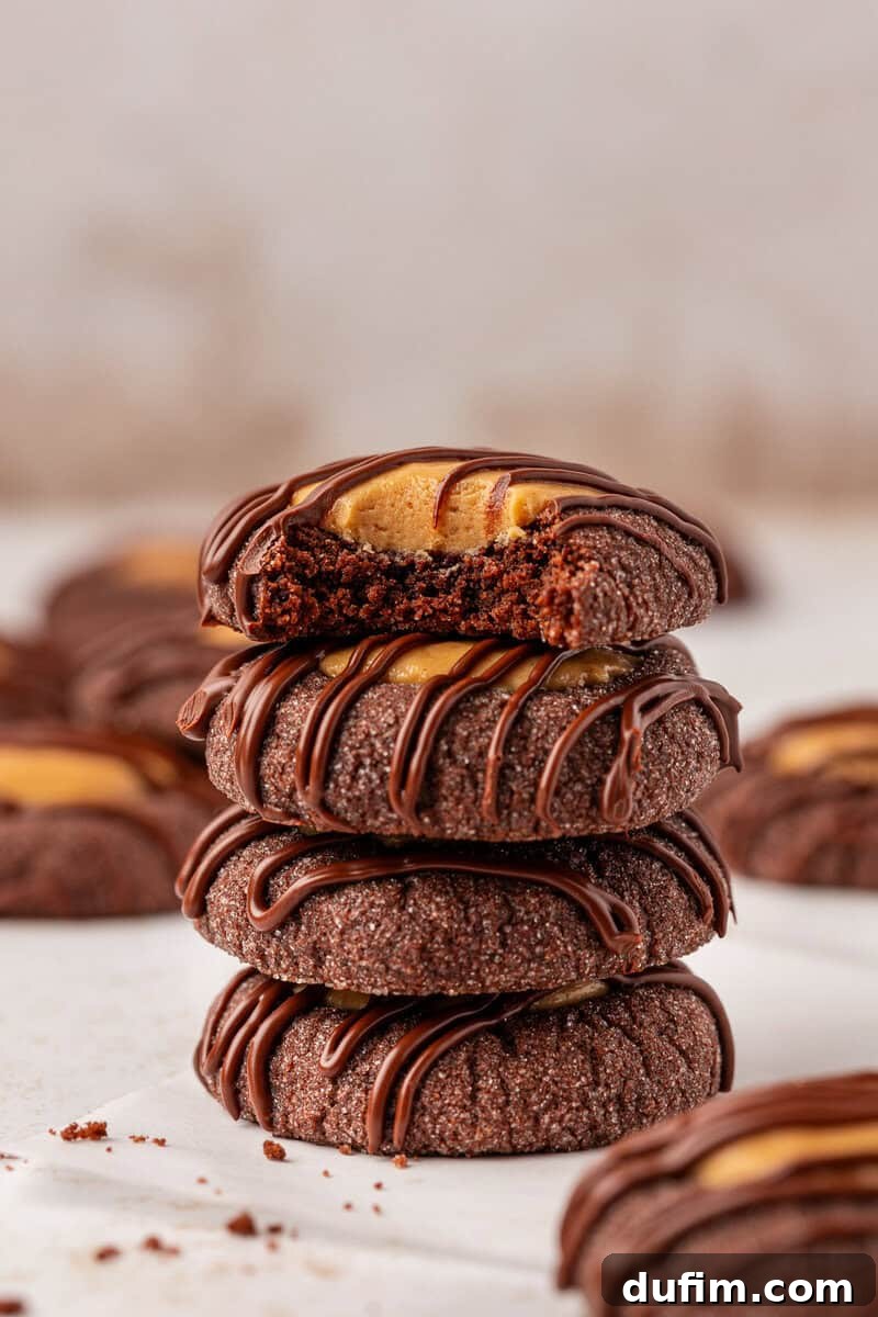 Irresistible Peanut Butter Chocolate Thumbprints 2 A stack of four chocolate cookies filled with peanut butter and drizzled with chocolate. The top cookie has a bite taken out, revealing the creamy peanut butter center. Crumbs are scattered on the white surface.