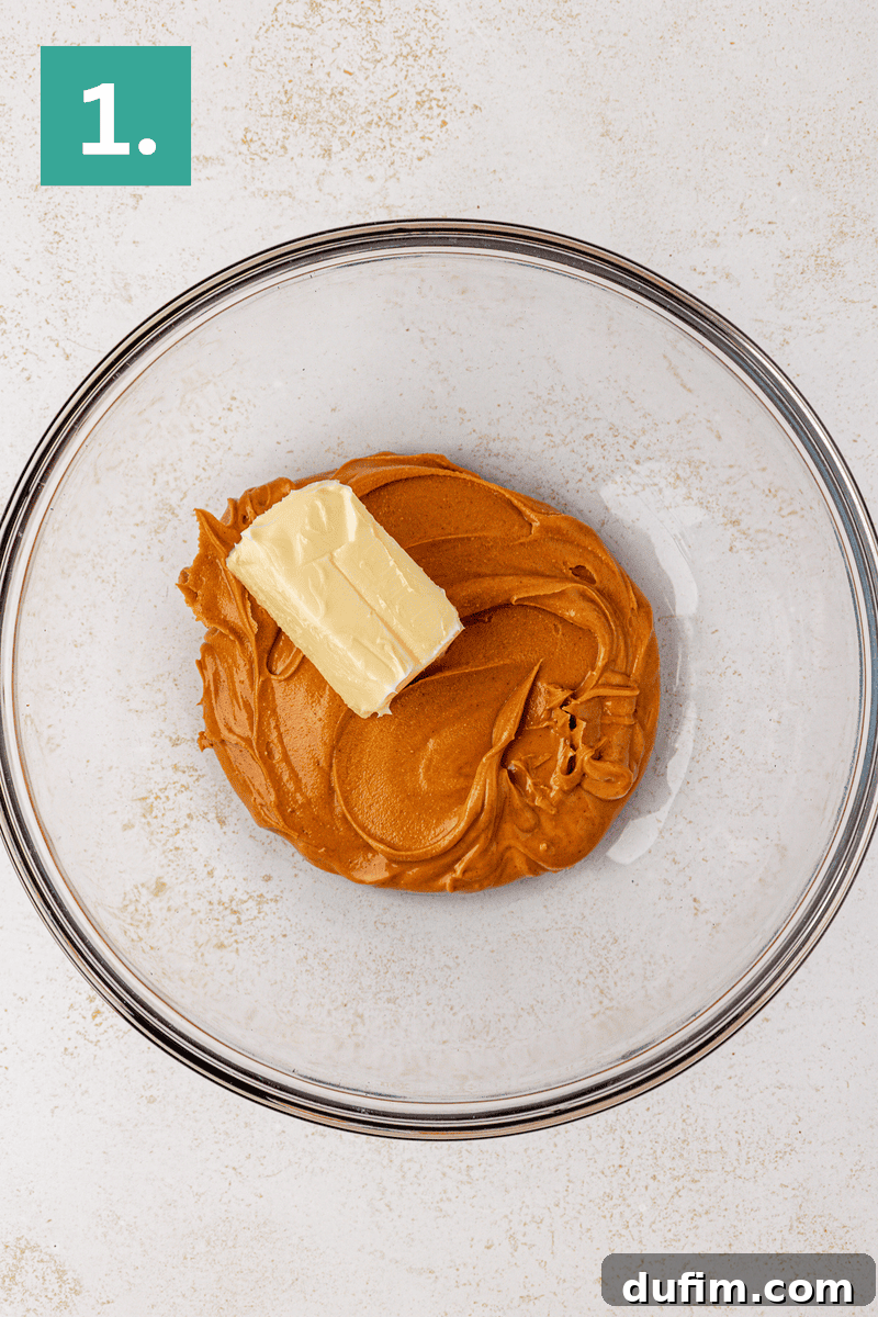 Irresistible Peanut Butter Chocolate Thumbprints 15 A glass mixing bowl containing creamy peanut butter and a chunk of butter. A green box with the number 1. is in the top left corner, indicating the first step of a recipe.