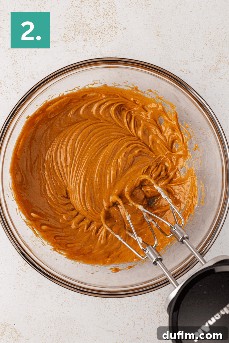 Irresistible Peanut Butter Chocolate Thumbprints 16 A glass bowl filled with creamy light brown batter being mixed with an electric hand mixer, shown from above. A green square with the number 2 appears in the top left corner.