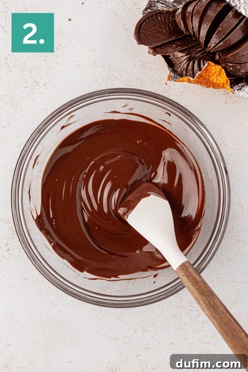 A glass bowl of melted chocolate being stirred with a white spatula. An opened chocolate orange sits in the top right corner. A green square with the number 2 is in the top left corner.